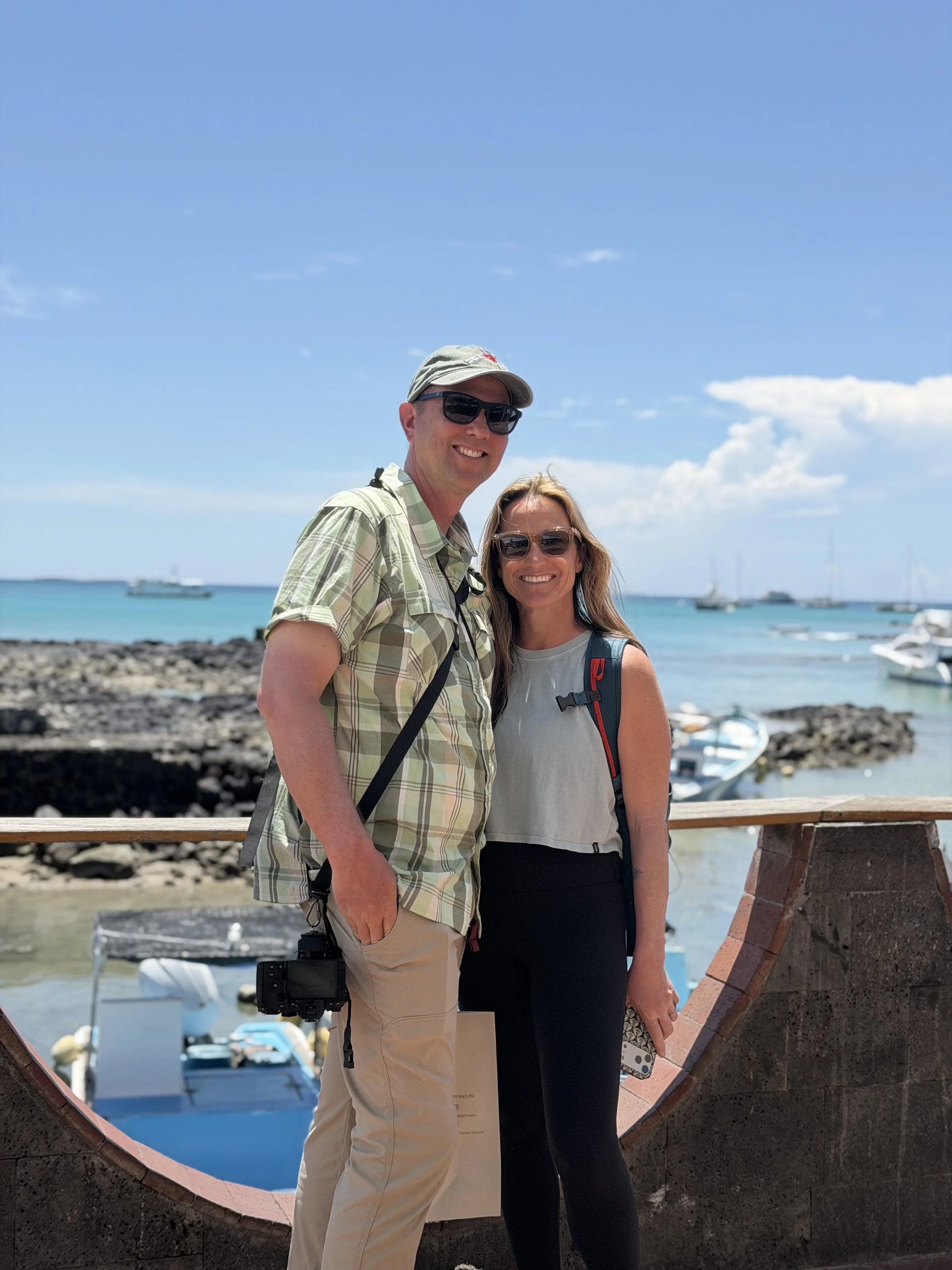 A man and woman smiling and wearing sunglasses, standing outdoors in Galapagos on a sunny day near the ocean, with boats and a rocky shoreline in the background.