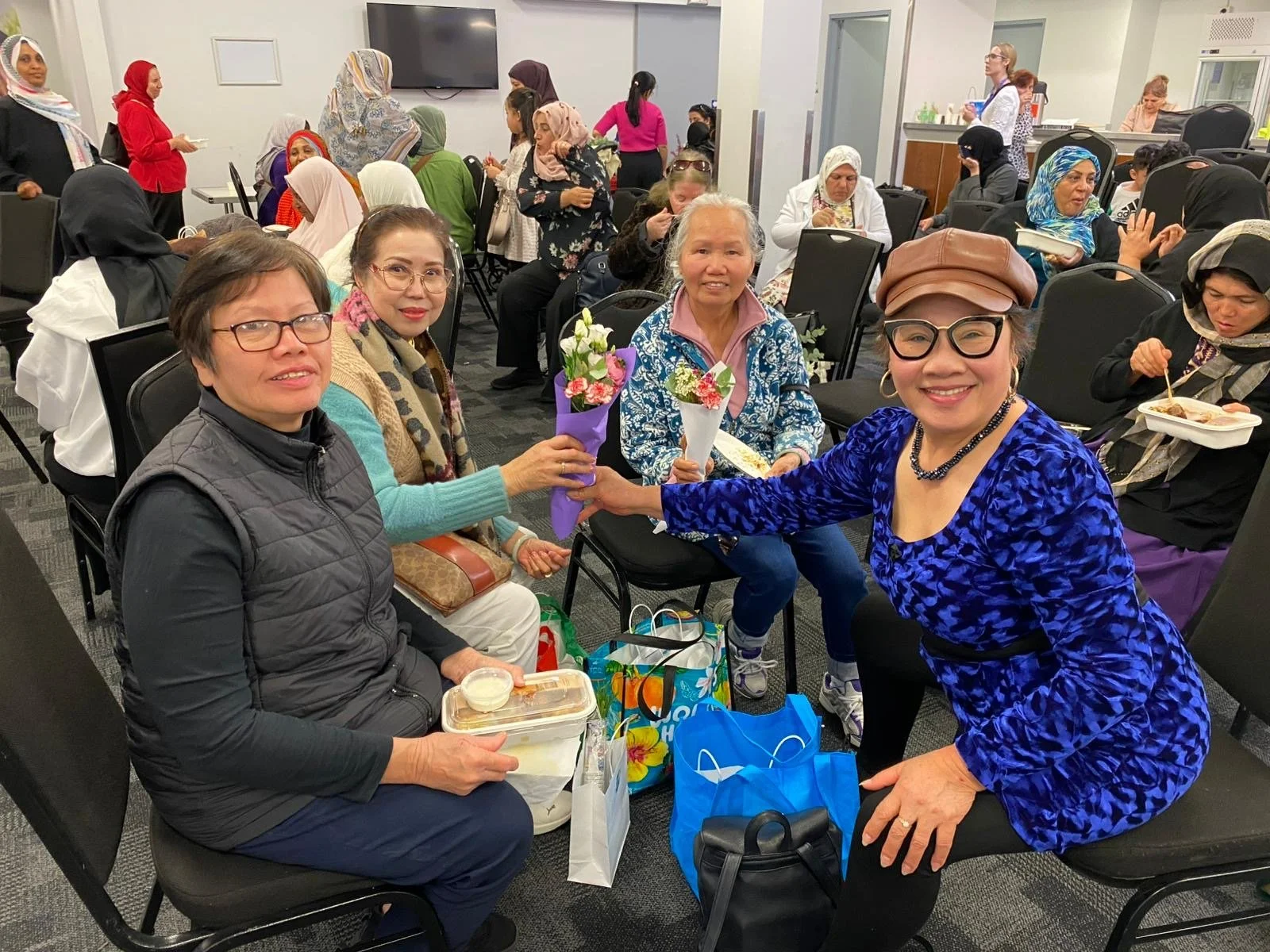 A group of diverse women gathered in a room, some sitting and some standing, with a woman in a blue dress and glasses exchanging flowers with a woman in a gray vest, while others enjoy food and conversation in the background.