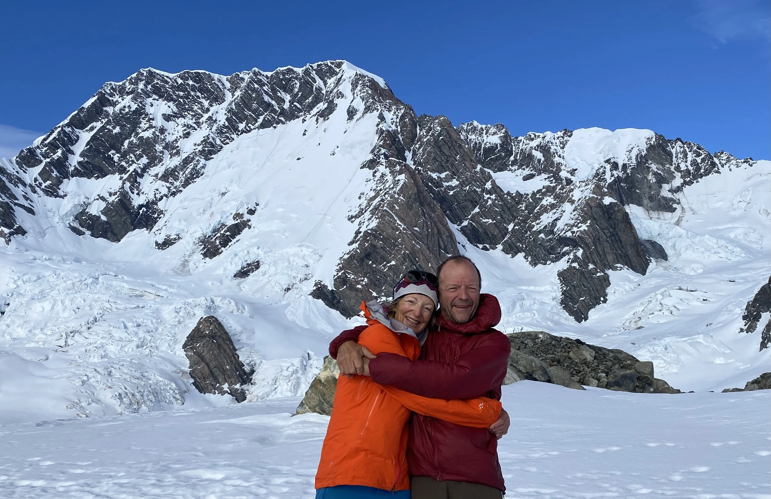 Lydia and Dean smiling and hugging in a snowy mountainous landscape. There are snow-covered mountains with rocky peaks in the background and a clear blue sky.