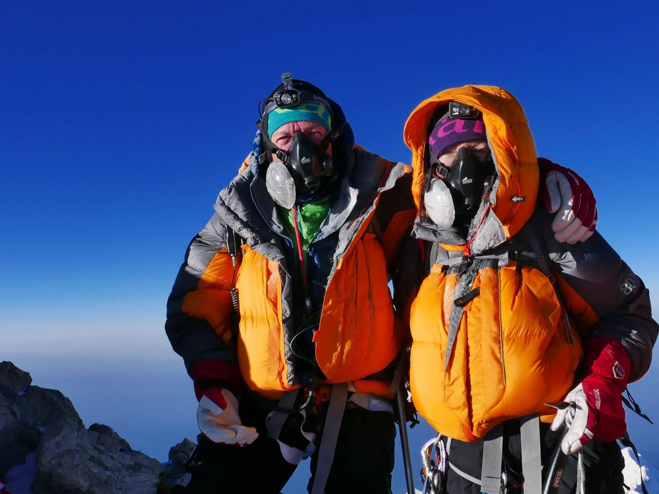 Lydia and Dean in winter gear with headlamps, standing on a rocky mountain peak under a clear blue sky.
