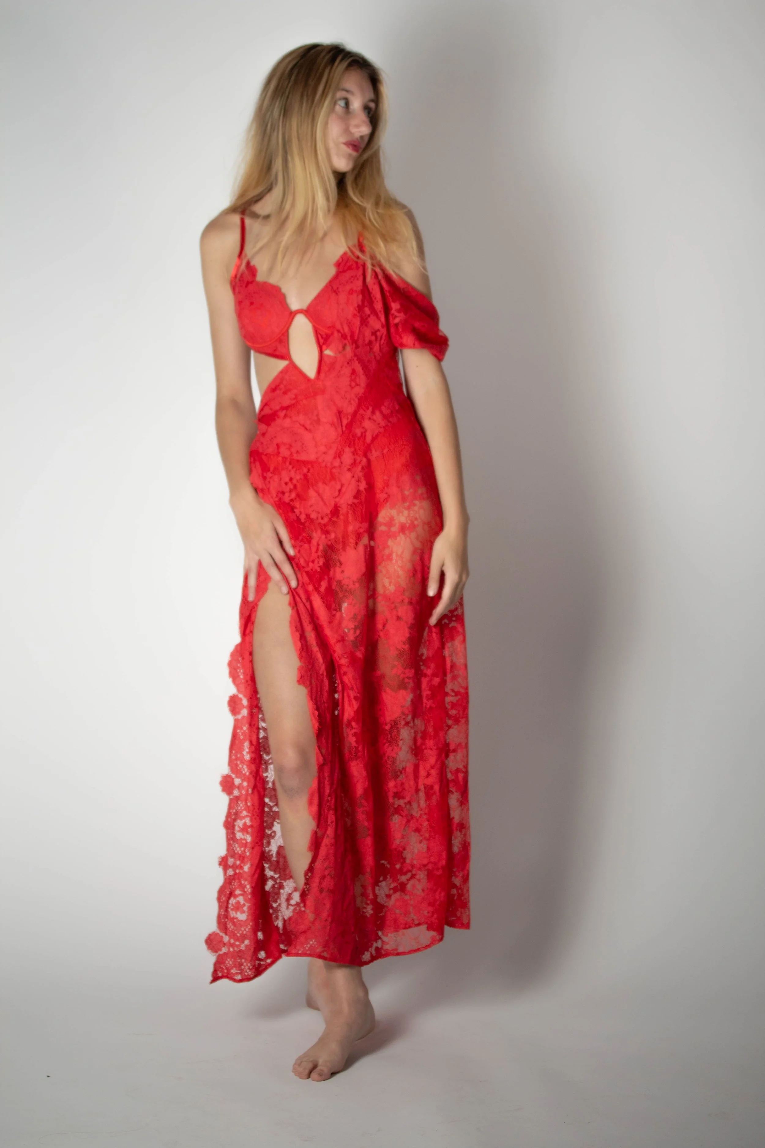 A woman in a red lace dress with a slit, standing barefoot against a plain wall.