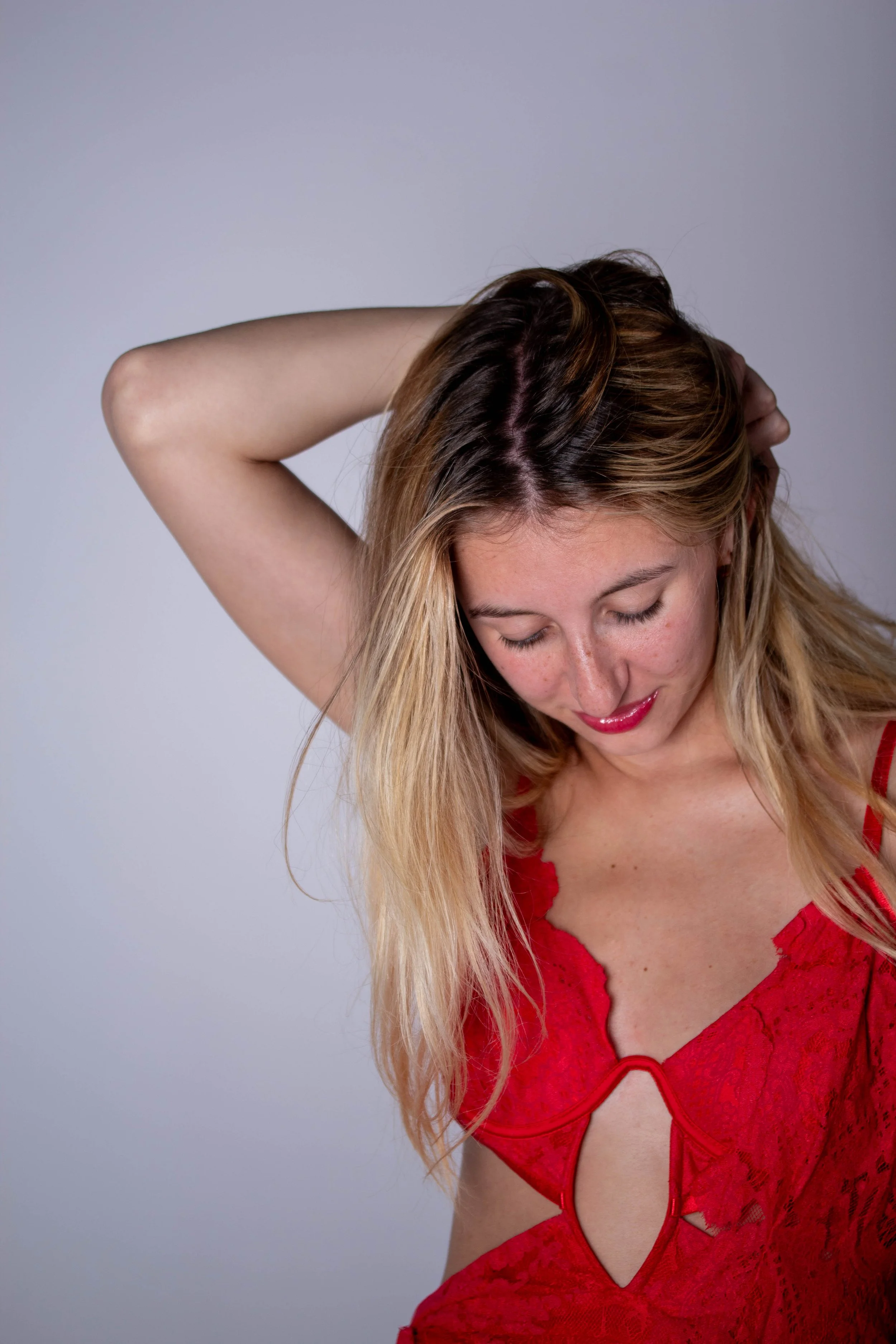 A woman with long blonde hair wearing a red lace dress, looking down and touching her hair while standing against a plain gray background.