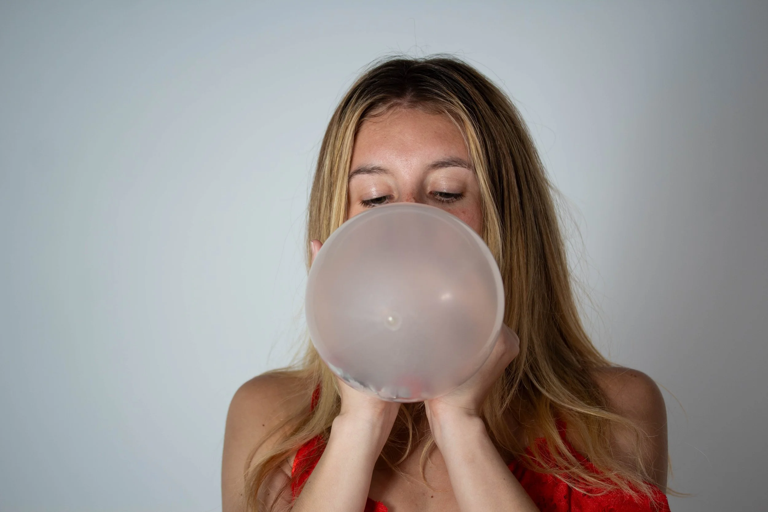 Woman with long blonde hair wearing a red dress, blowing up a balloon.