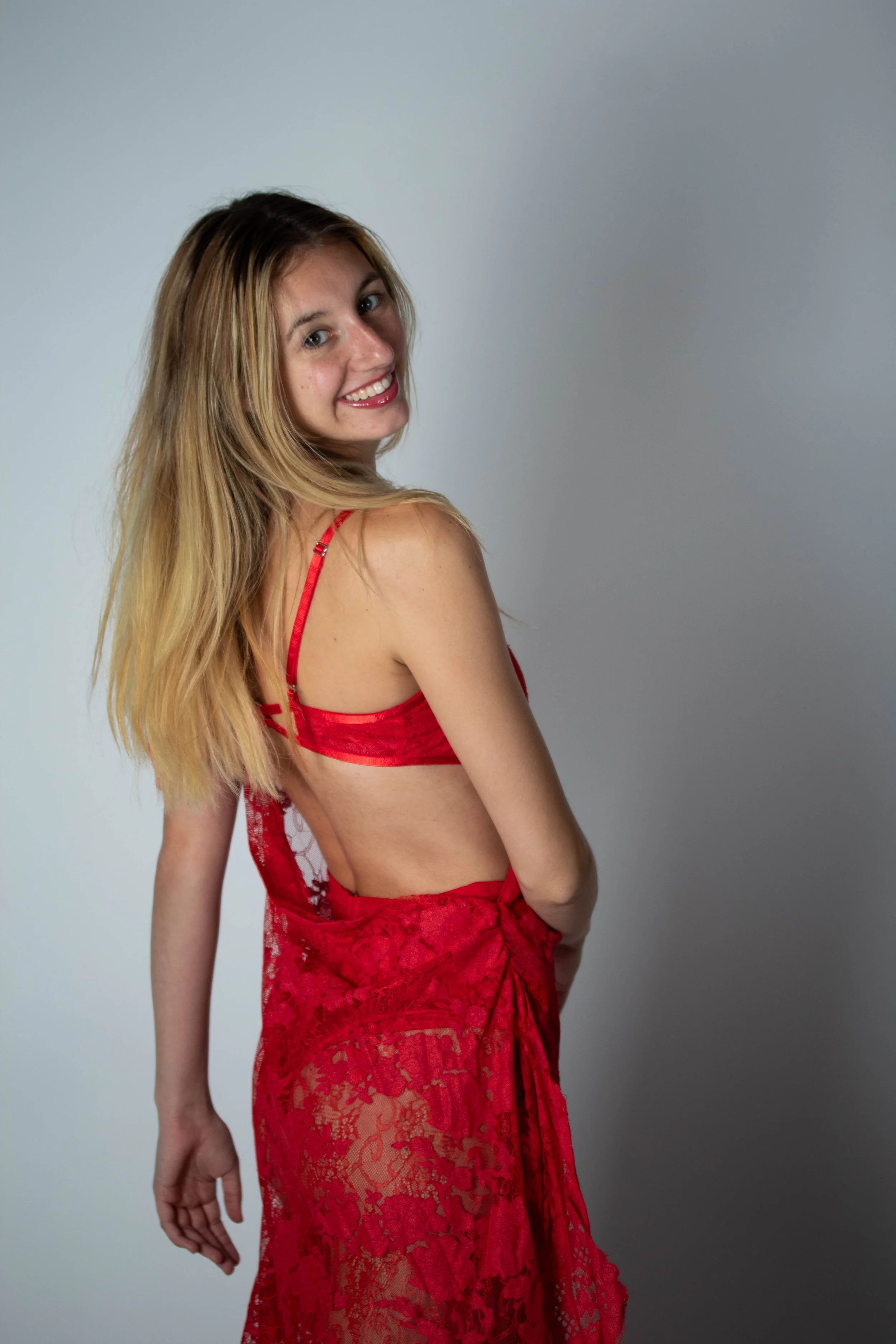 A woman with long blonde hair wearing a red dress, smiling and turning her head over her shoulder against a plain gray background.
