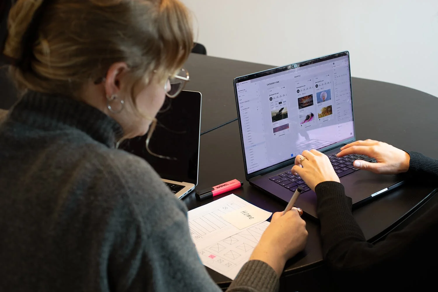 Two people are working at a table with a laptop and drawing paper, on a black table in a meeting room.