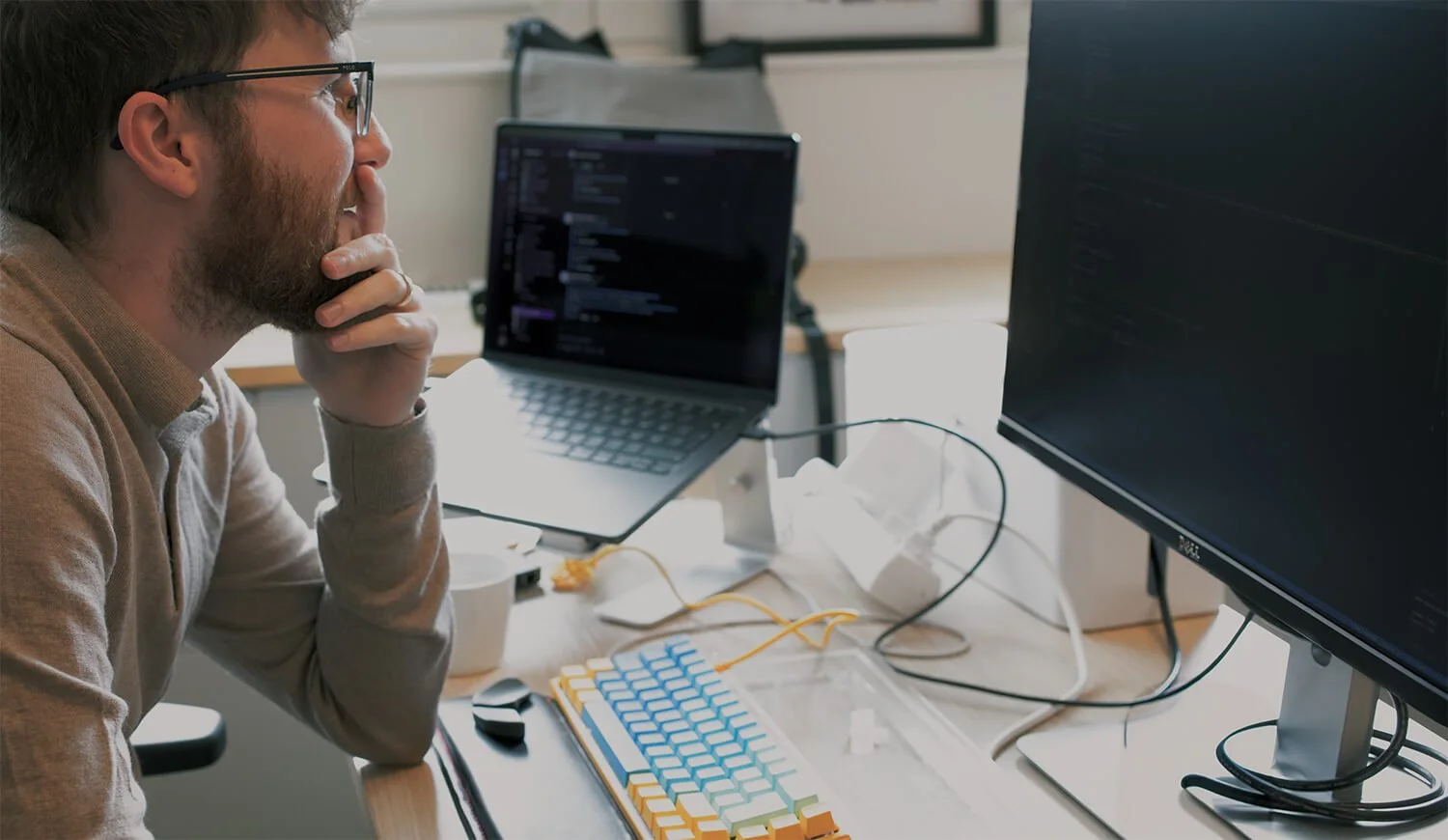 Man with glasses and beard looking at a screen in an office environment, with two laptops and a keyboard on the desk.
