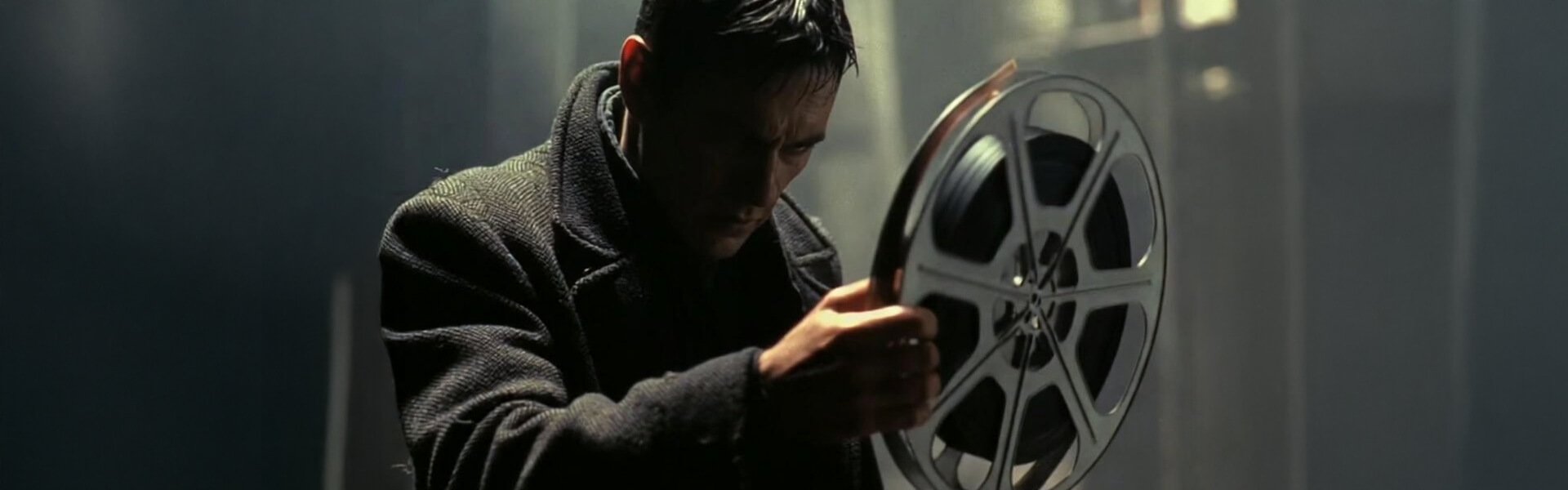 Man in dark coat gazing thoughtfully at a large film reel he holds in both hands, in low light.