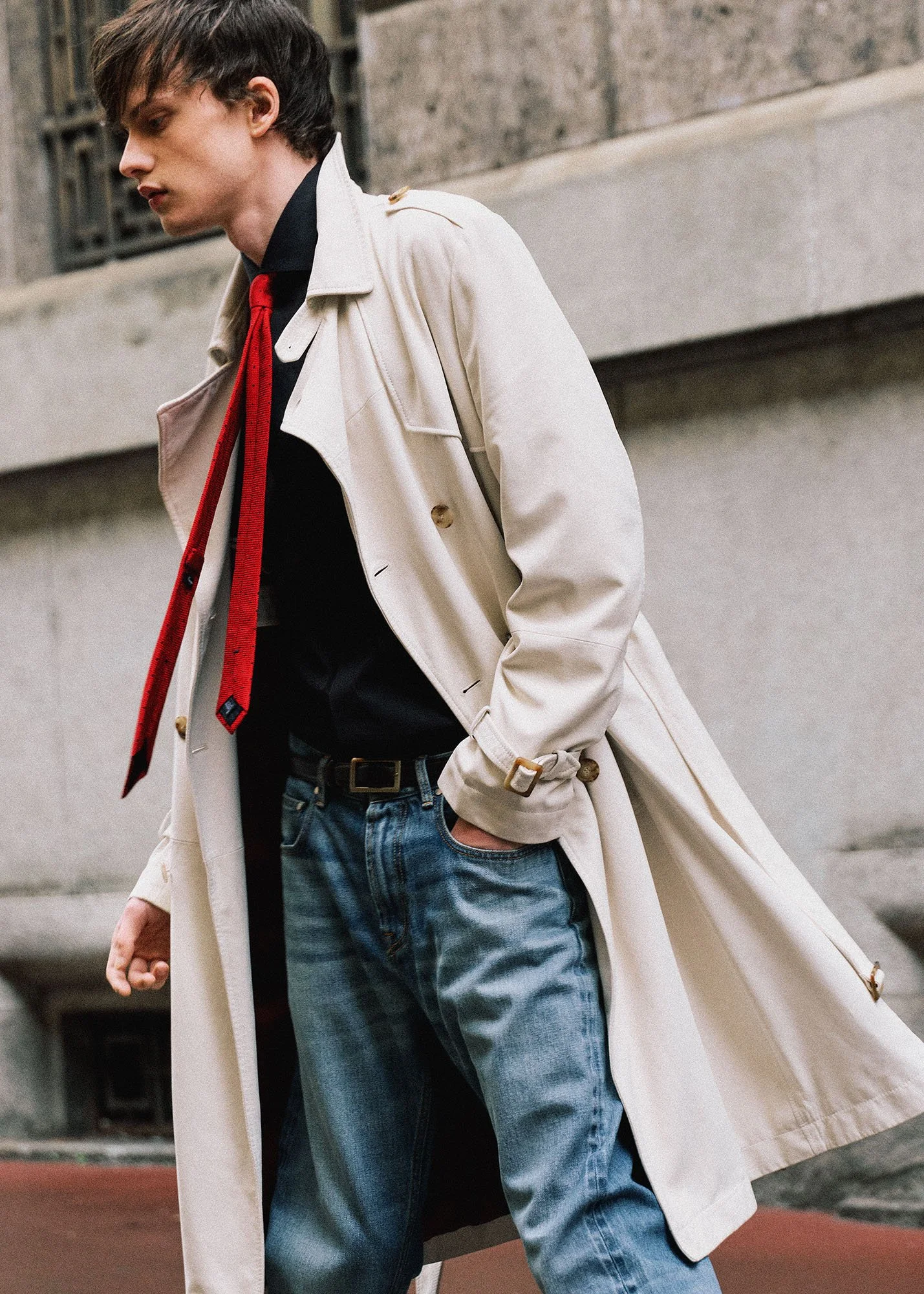 A young man walking outdoors, wearing a white trench coat, black shirt, red tie, and blue jeans, with his hand in his pocket.