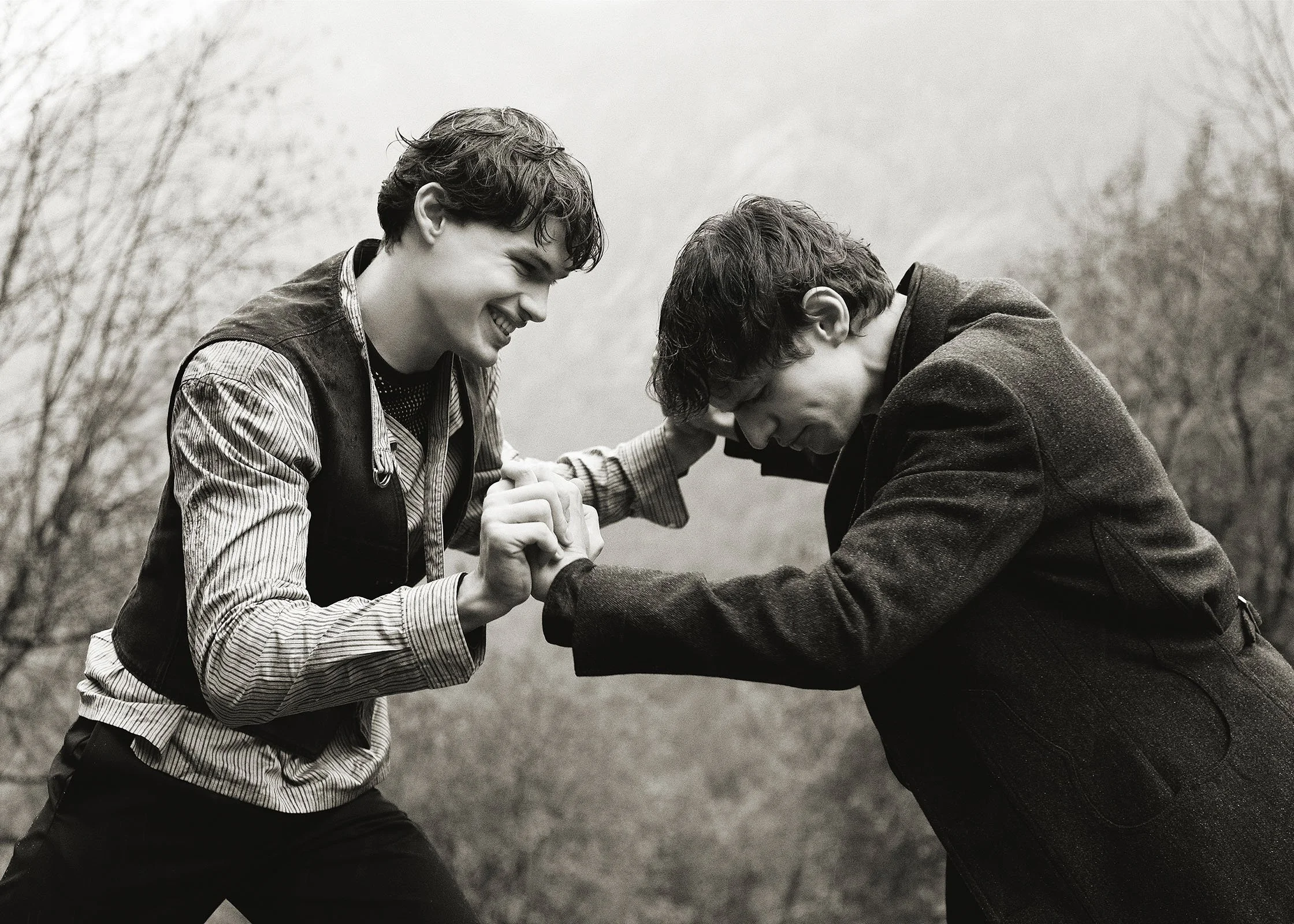 Two young men engaging in an arm wrestling match outdoors, smiling and leaning into each other, with trees in the background.