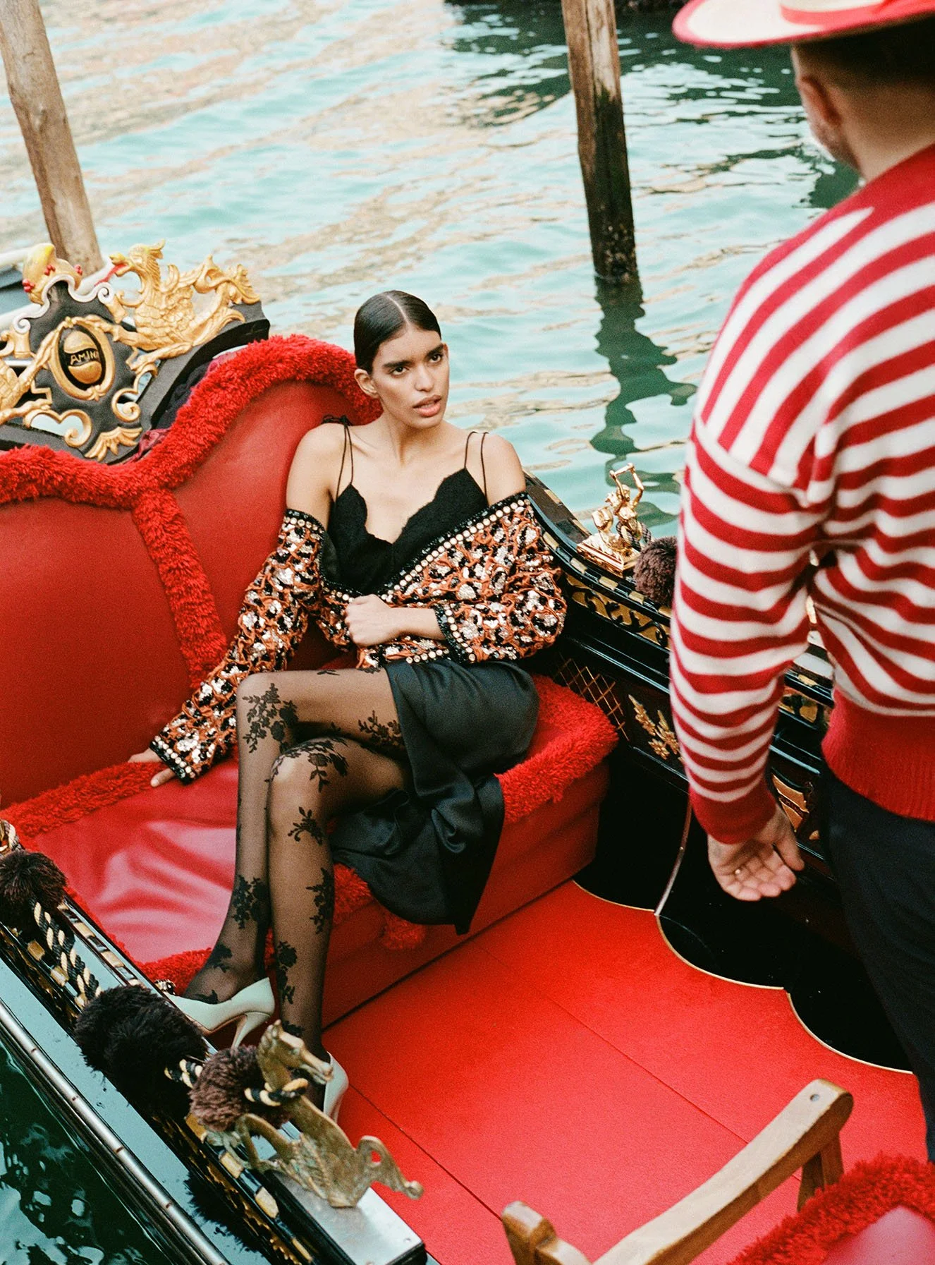 A woman sitting on an ornate red and gold gondola, partially submerged in water, wearing black lace tights, a black satin skirt, and an embroidered jacket, while a man in a red and white striped sweater stands nearby.