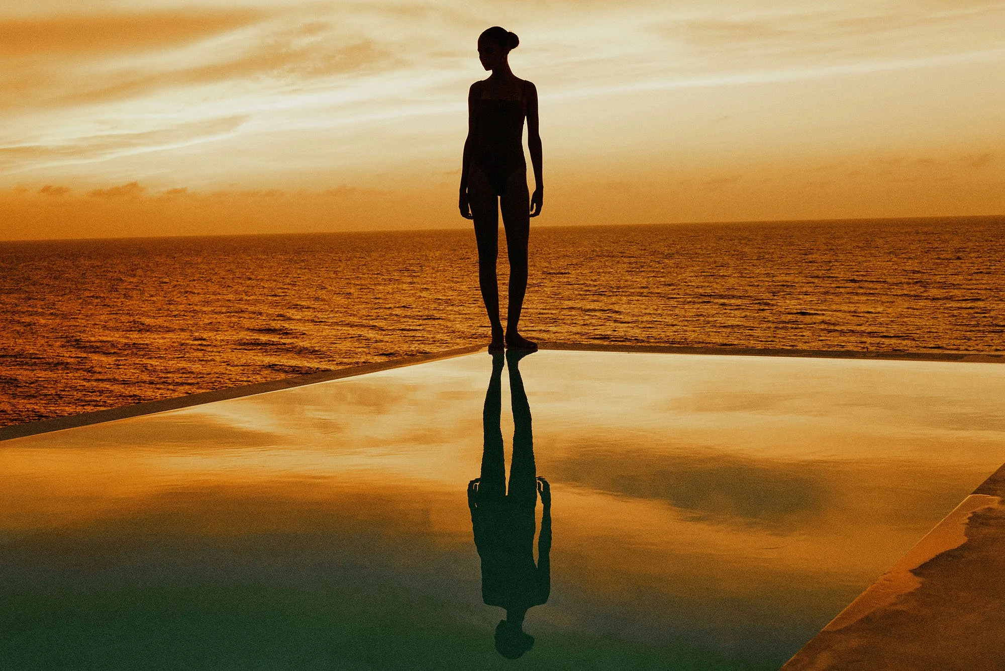 Silhouette of a woman standing on the edge of a reflective surface at sunset, with the ocean in the background and the girl's reflection visible in the surface.