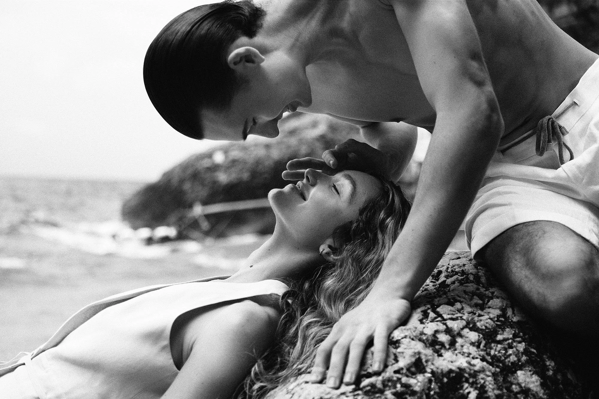 A black and white photo of a shirtless man leaning over a woman lying on a rock at the beach, with the ocean and a distant rocky island in the background. The man is gently touching the woman's forehead while she smiles with her eyes closed.