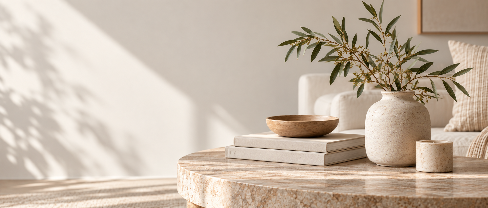 Sunlit styled vignette of a round stone coffee table with books, a timber bowl and olive branches in a cream vase