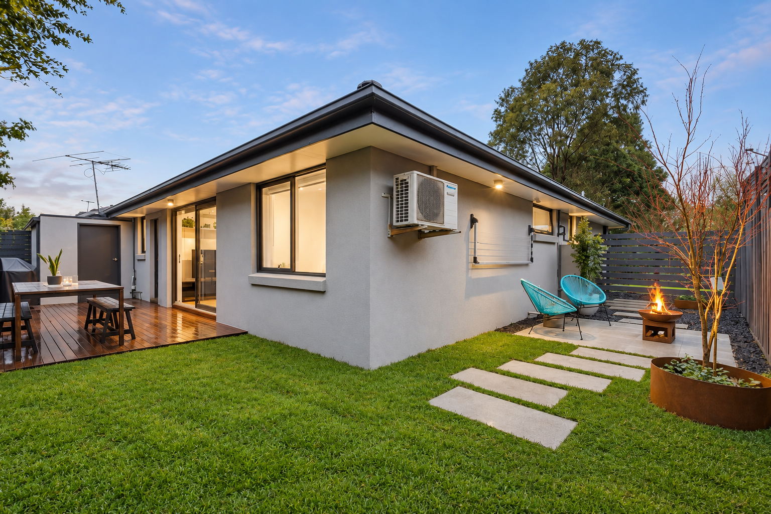 Centre project rear exterior: grey rendered home with lawn, stepping-stone path, timber deck and fire pit at dusk