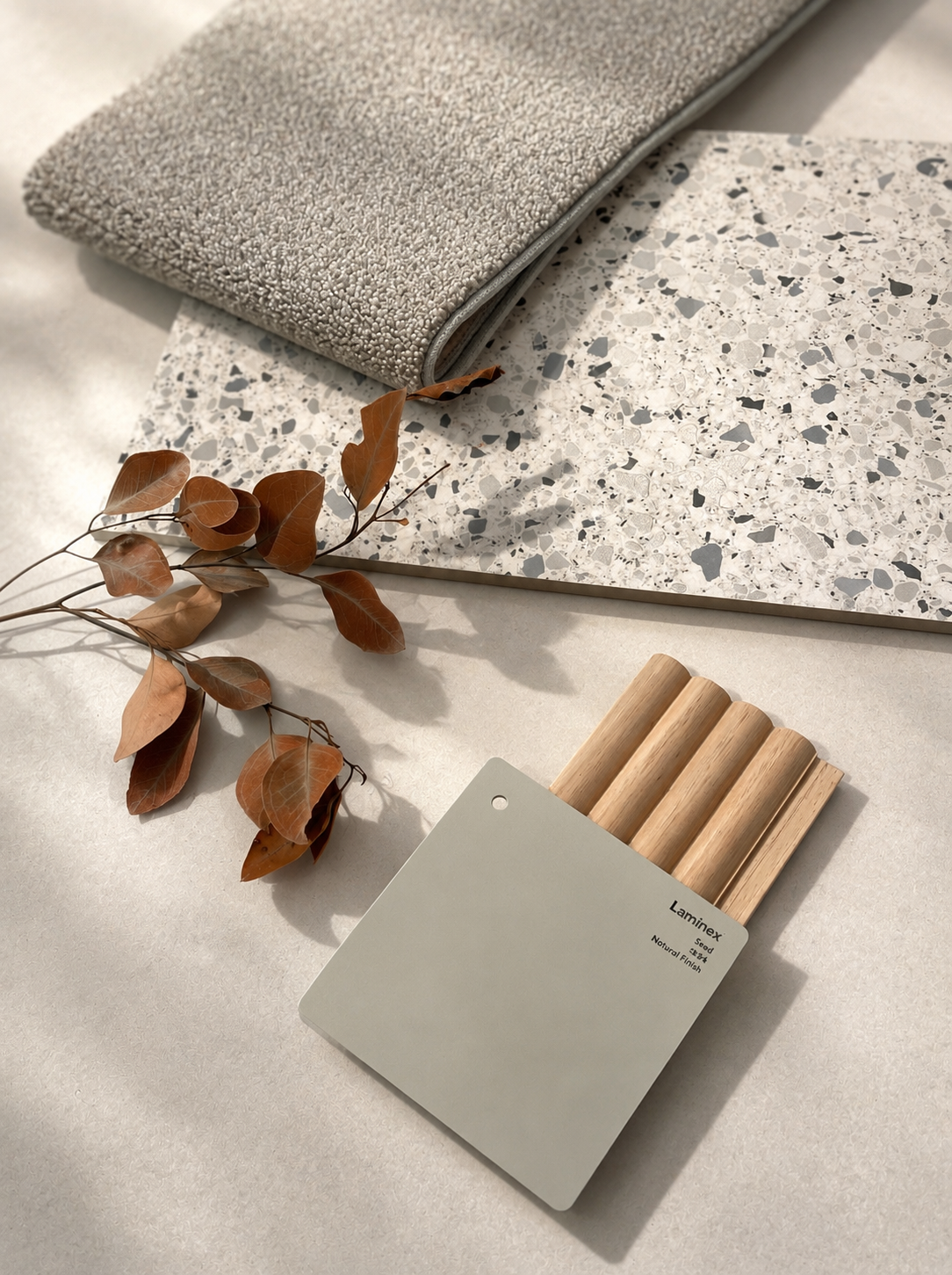 Flatlay of grey carpet, terrazzo, sage laminate swatch and timber battens with autumn eucalyptus leaves