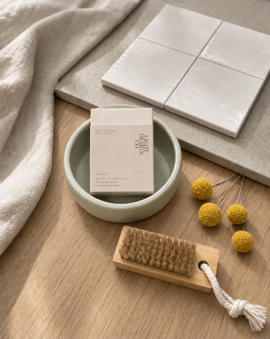 Light Design Co. cleanse bar in a sage ceramic bowl with glazed white tiles, a timber nail brush, linen towel and yellow billy buttons