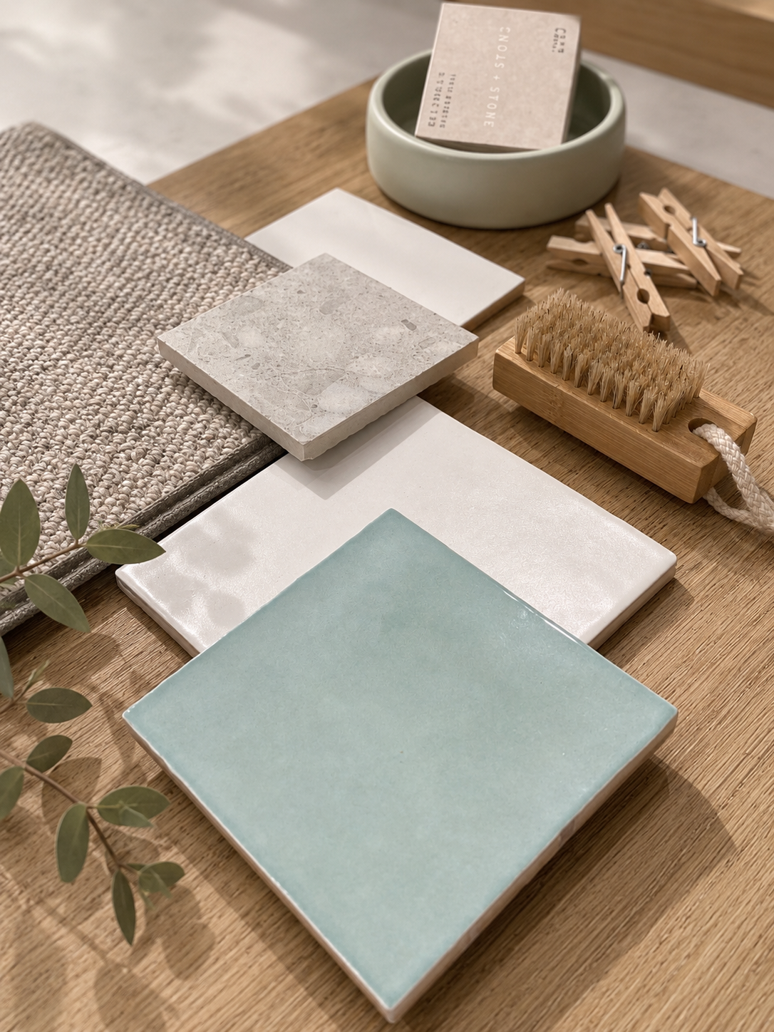 Flatlay of a sea-green glazed tile, grey carpet, stone samples, sage bowl, timber brush and eucalyptus on oak