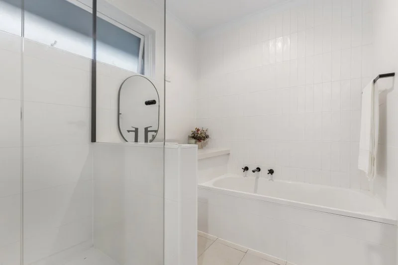 Centre project bathroom with white tiles, frameless glass shower, built-in bath and black tapware