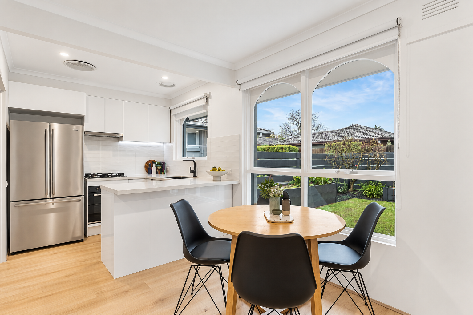 Centre project bright white kitchen and dining nook with round timber table, black chairs and garden views