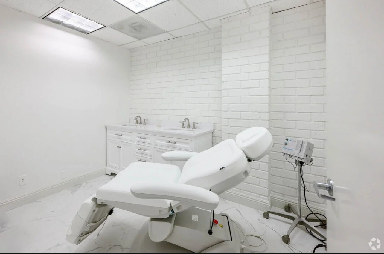A medical examination or treatment room with a white adjustable examination chair, a white brick wall, a sink with two faucets, white cabinets, and medical equipment on a stand.