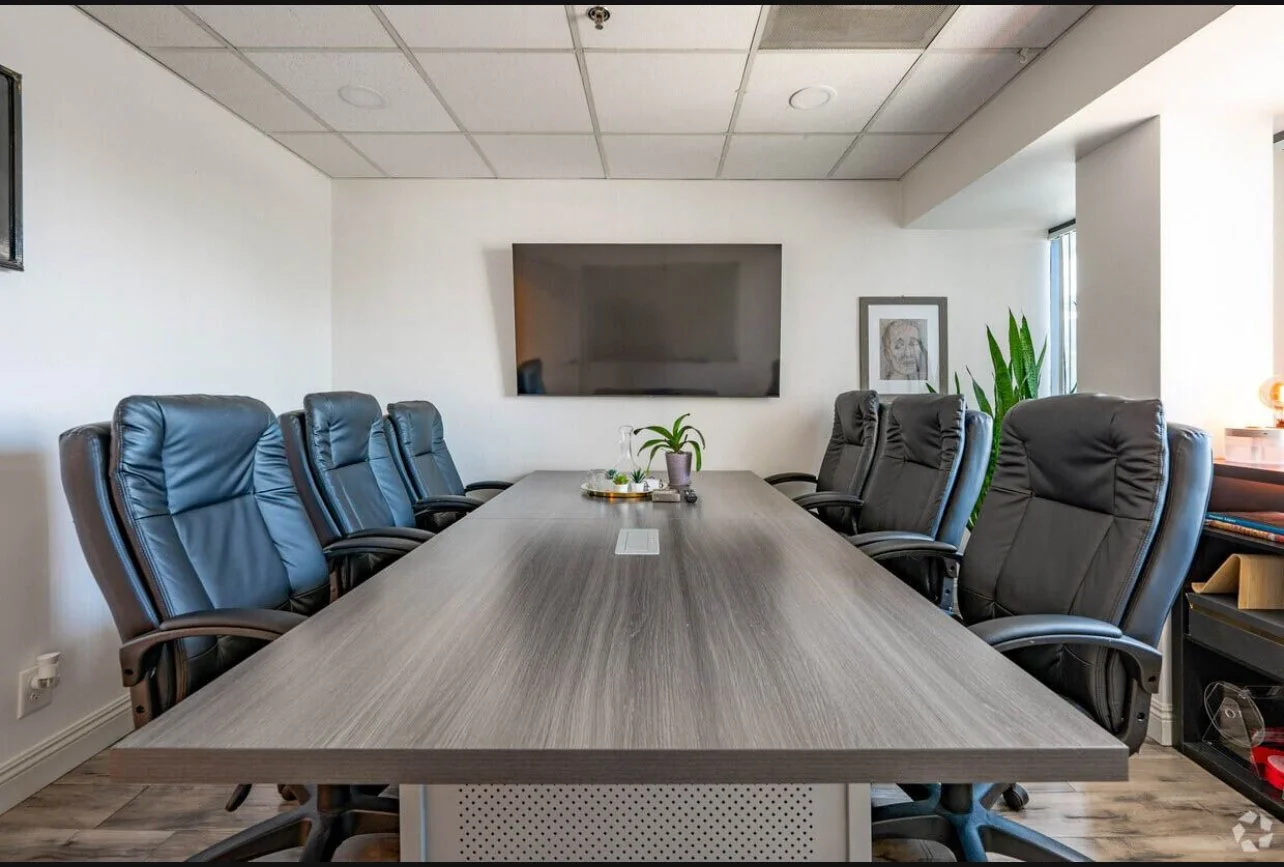 Empty conference room with a large wooden table surrounded by black leather office chairs, a wall-mounted television, a framed portrait, and a plant in the corner.
