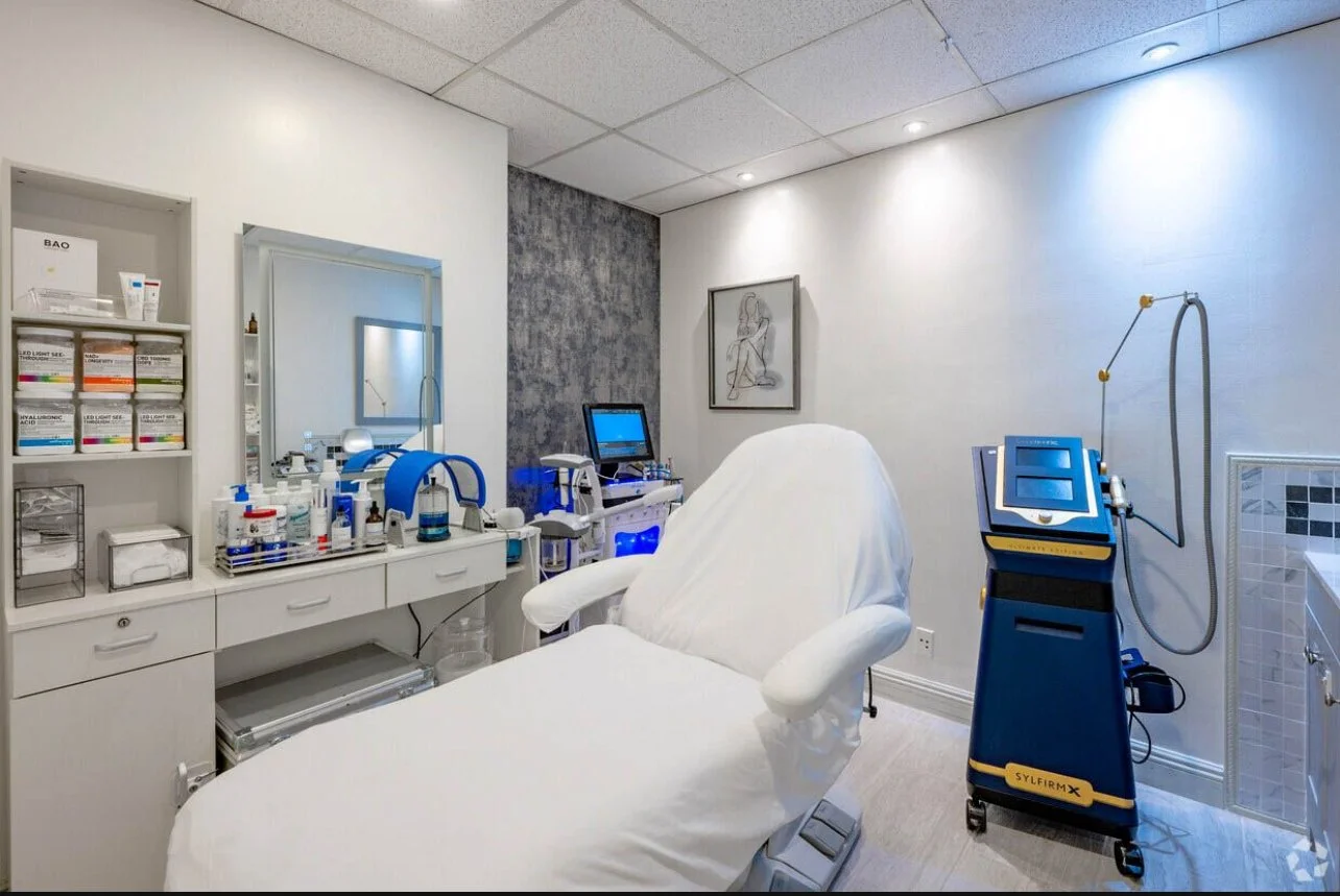 A medical treatment room with a white reclining medical chair, medical equipment, and supplies on a countertop. There is a mirror on the wall, a framed artwork, and medical devices including a blue and black machine, all under a ceiling with recessed