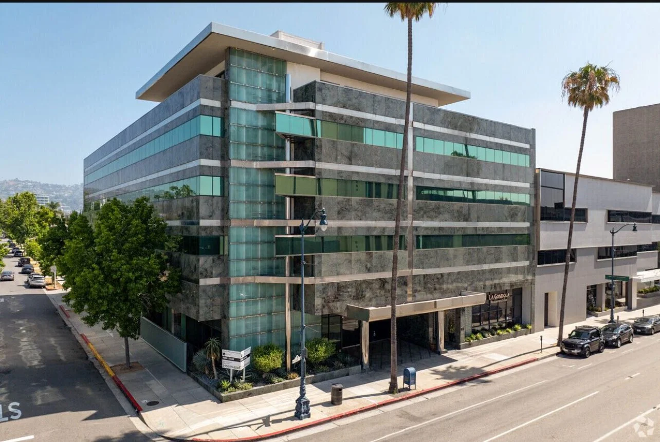 Modern office building with glass and gray concrete facade on a sunny day, surrounded by palm trees and parked cars.