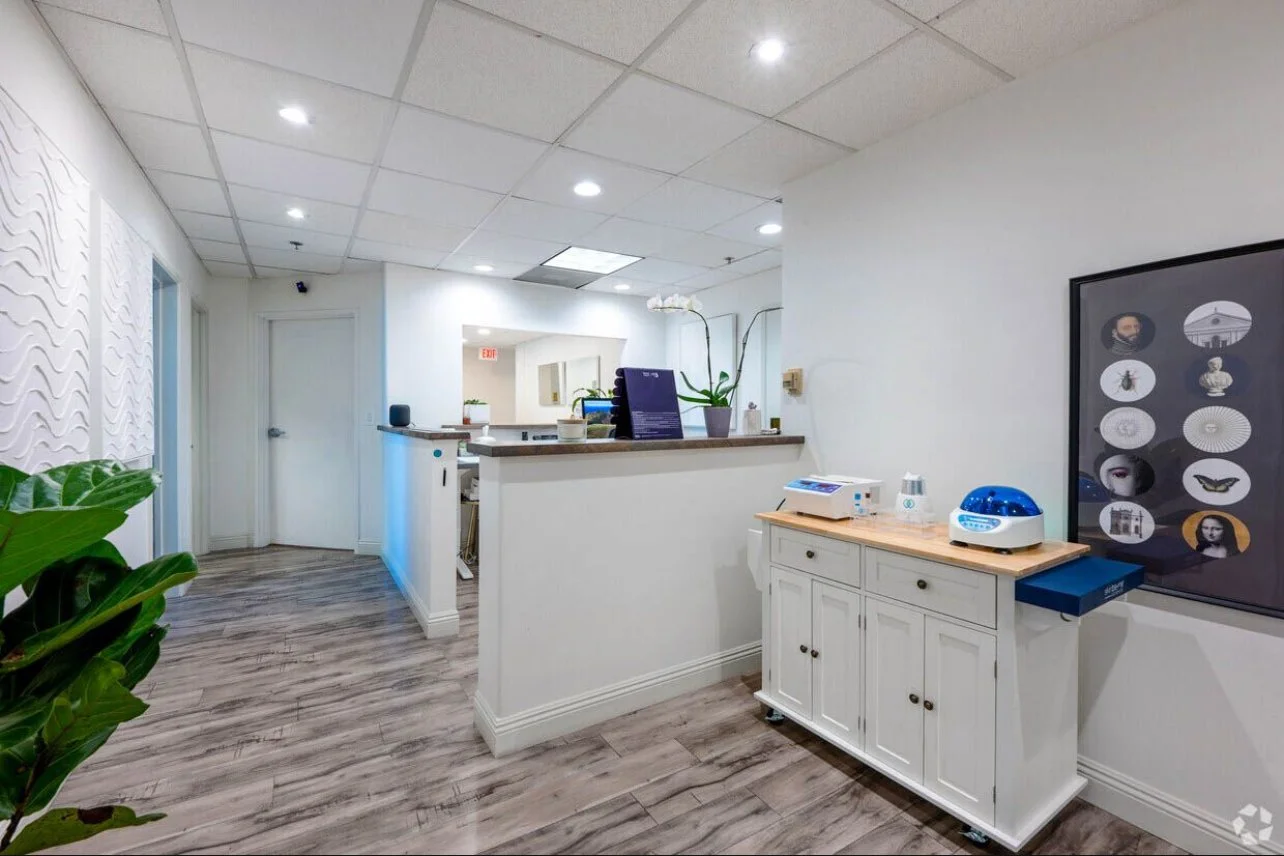 Interior of a modern medical or dental office reception area with white walls, wood flooring, a white cabinet on wheels, medical or dental equipment on top, and a framed artwork on the wall.