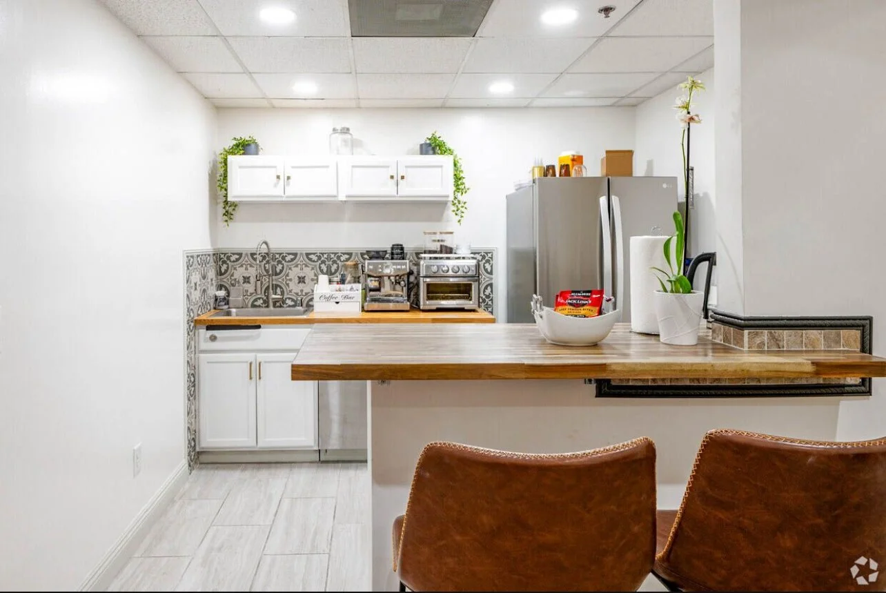 A small, modern kitchen with white cabinets, patterned backsplash, and wooden countertops. There is a refrigerator, a stove, and a coffee maker. A bowl with snacks and plants are on the counter, with two brown chairs in front.