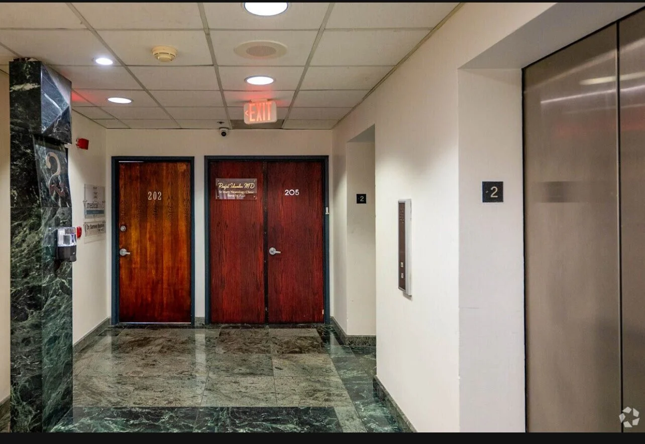 Hallway with an elevator on the right, two wooden doors labeled 202 and 205, a wall-mounted intercom, an exit sign on the ceiling, and an elevator button panel.