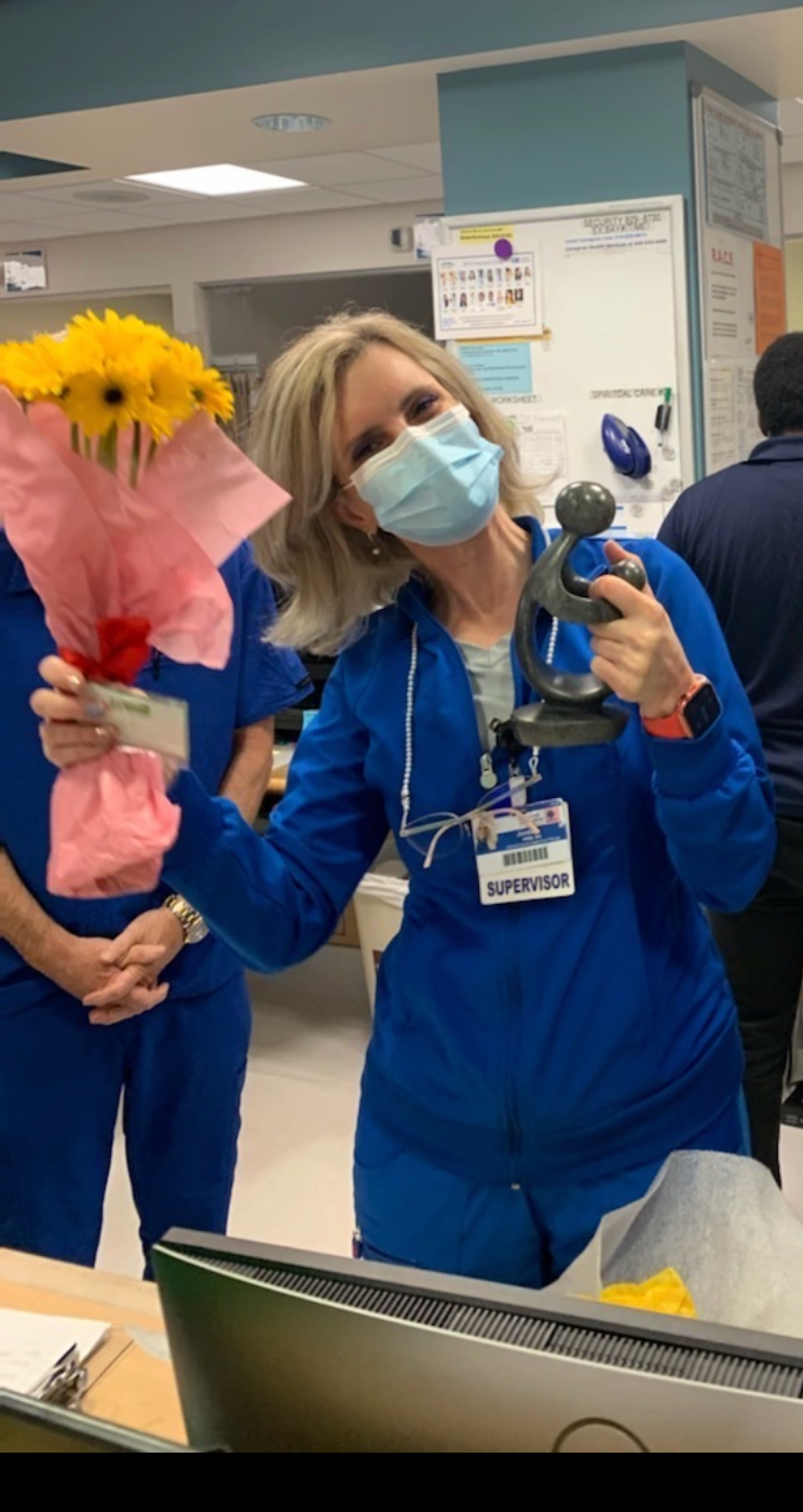 A woman in a blue uniform with a 'Supervisor' badge and a face mask is smiling and holding a bouquet of yellow flowers wrapped in pink paper and a small anchor figurine. She is in a hospital or medical facility, with other people and medical notices visible in the background.