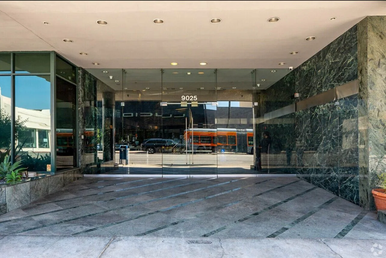 View of a modern building entrance with glass doors, green marble walls, and plants outside. Reflection of passing streetcars and cars in the glass.