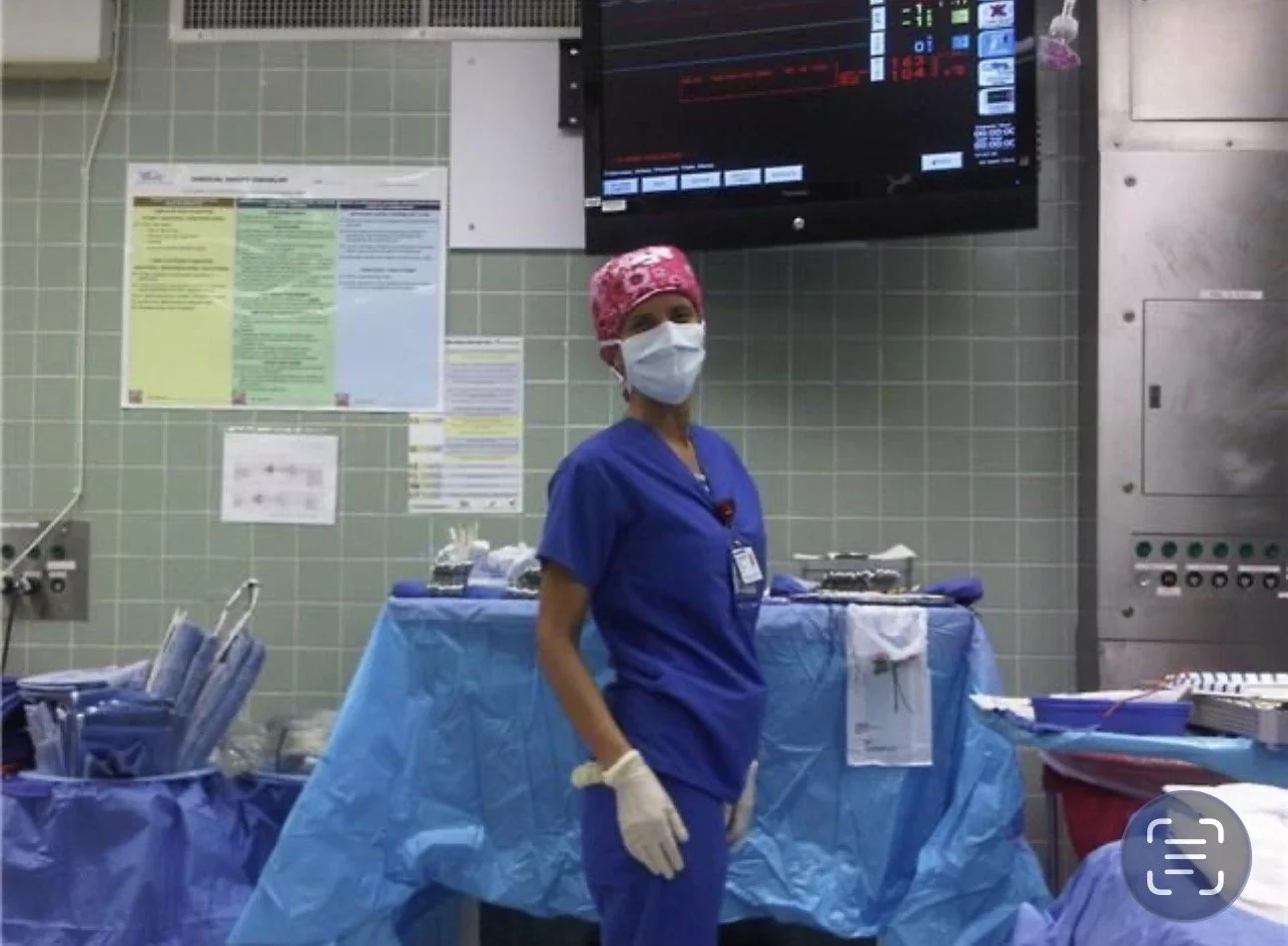 A healthcare worker in blue scrubs, wearing a pink patterned surgical cap, white gloves, and a face mask, standing in a hospital operating room with medical equipment and supplies around.