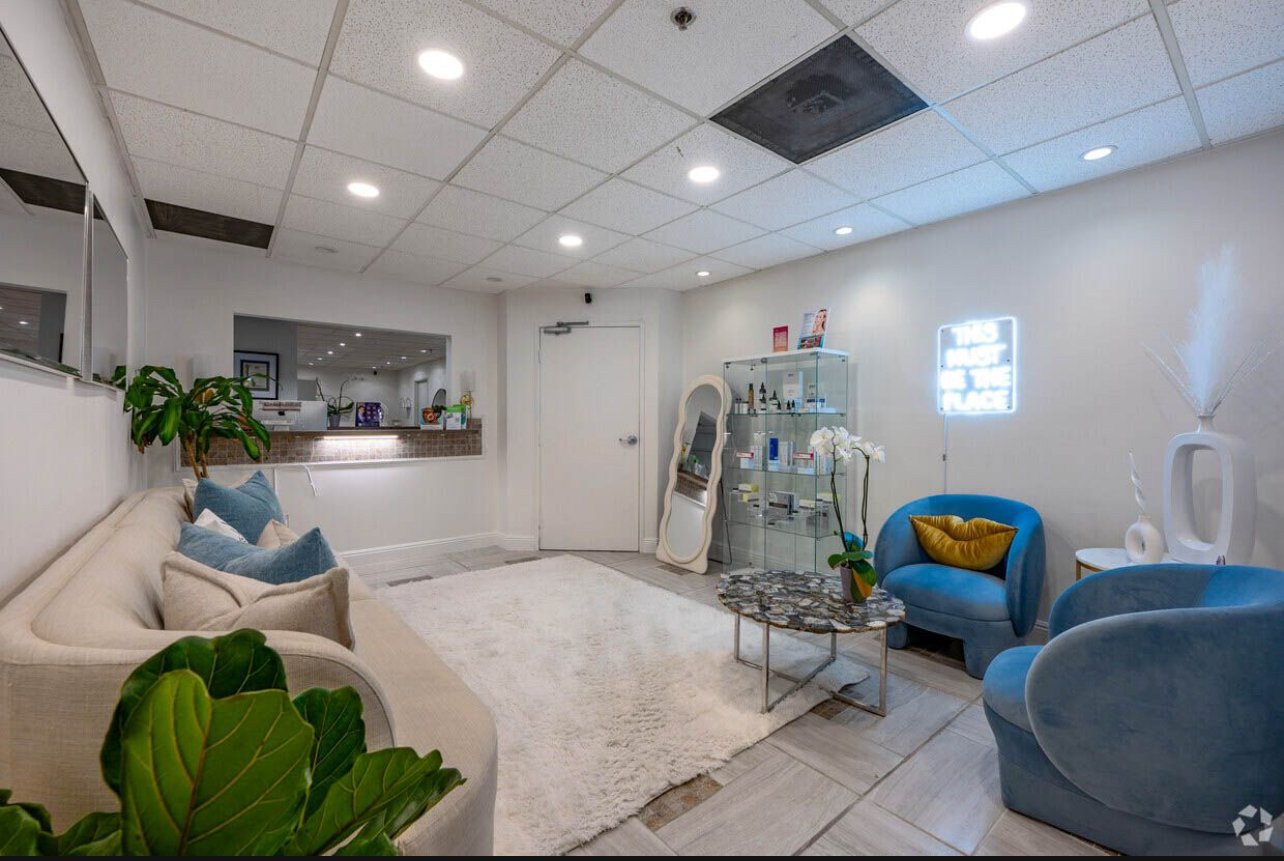 Modern reception or waiting area with a beige sofa decorated with cushions, a blue armchair with a yellow pillow, a marble coffee table, a white vase with white flowers, a glass display cabinet with products, and neon wall art reading 'This is the pl