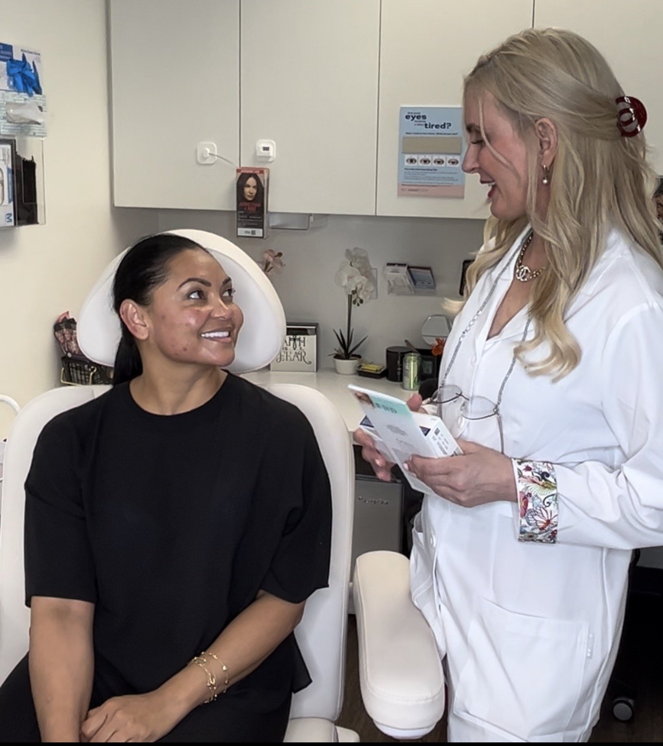 A woman with long dark hair sitting in a chair at a medical clinic, smiling and looking at a healthcare professional, a blonde woman in a white coat, who is holding a pamphlet or brochure and talking to her.