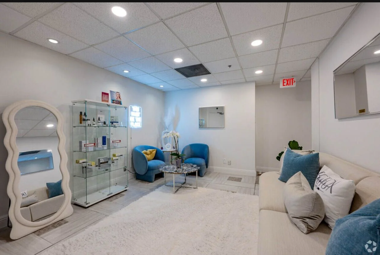 A modern waiting area with blue chairs, a white sofa with pillows, a glass display cabinet, a large mirror, and a white rug, with white walls and a ceiling with recessed lighting.