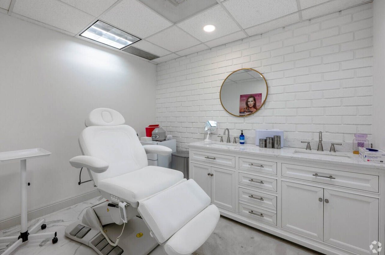 A medical examination room with a white reclining chair, white cabinetry, a mirror, and medical supplies visible.