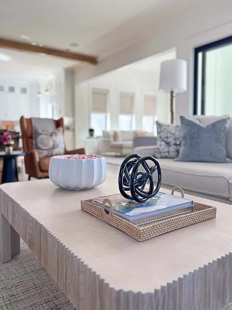 Living room with a beige coffee table featuring a decorative black sculpture, a wicker tray with books, and a white bowl. Background includes a couch with pillows, a floor lamp, and a window with blinds.