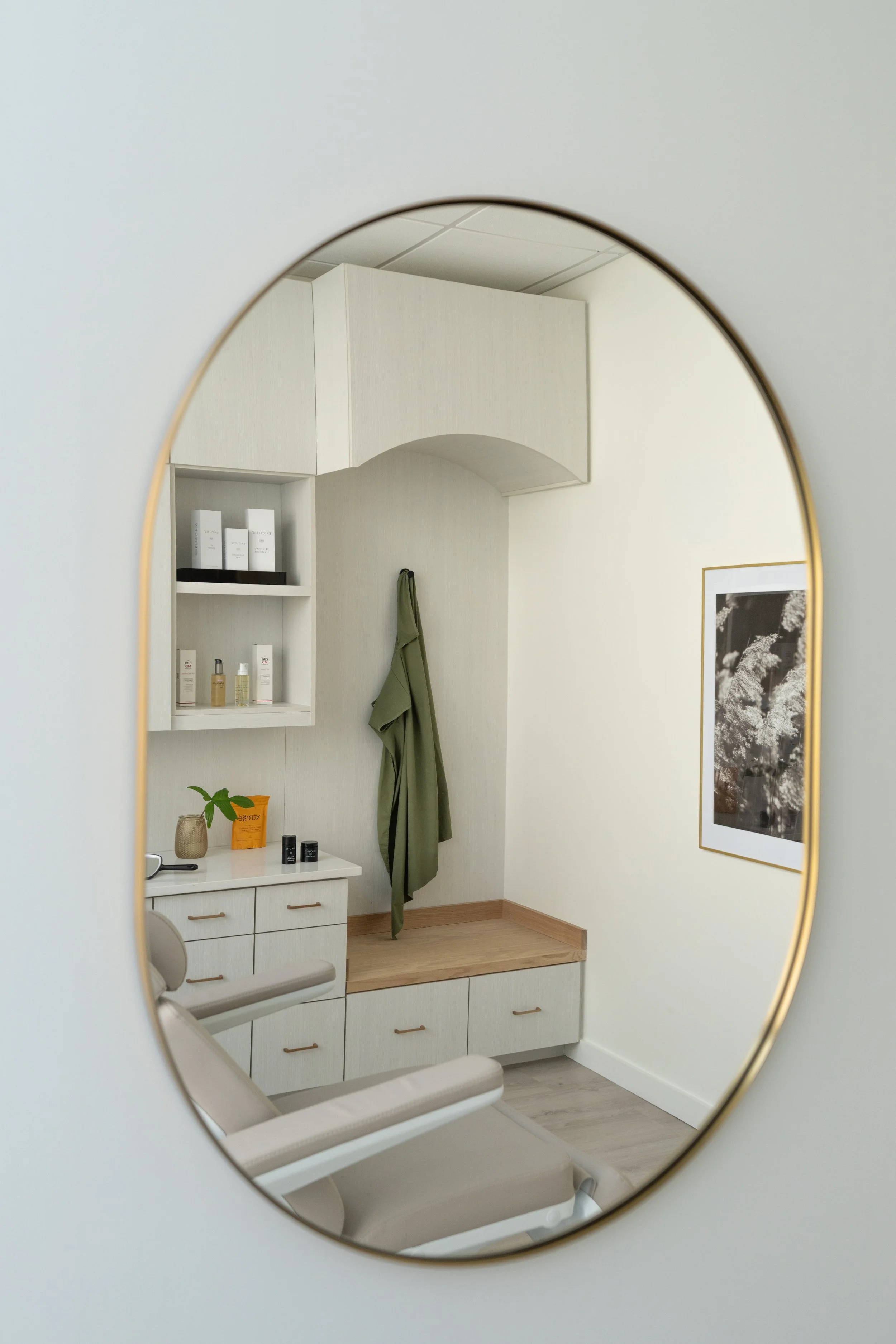 A reflection of a white medical or dental office room in a mirror, showing a tray table, a wall with shelves holding skincare or medical products, a green robe hanging, and a framed black-and-white photograph of flowers.