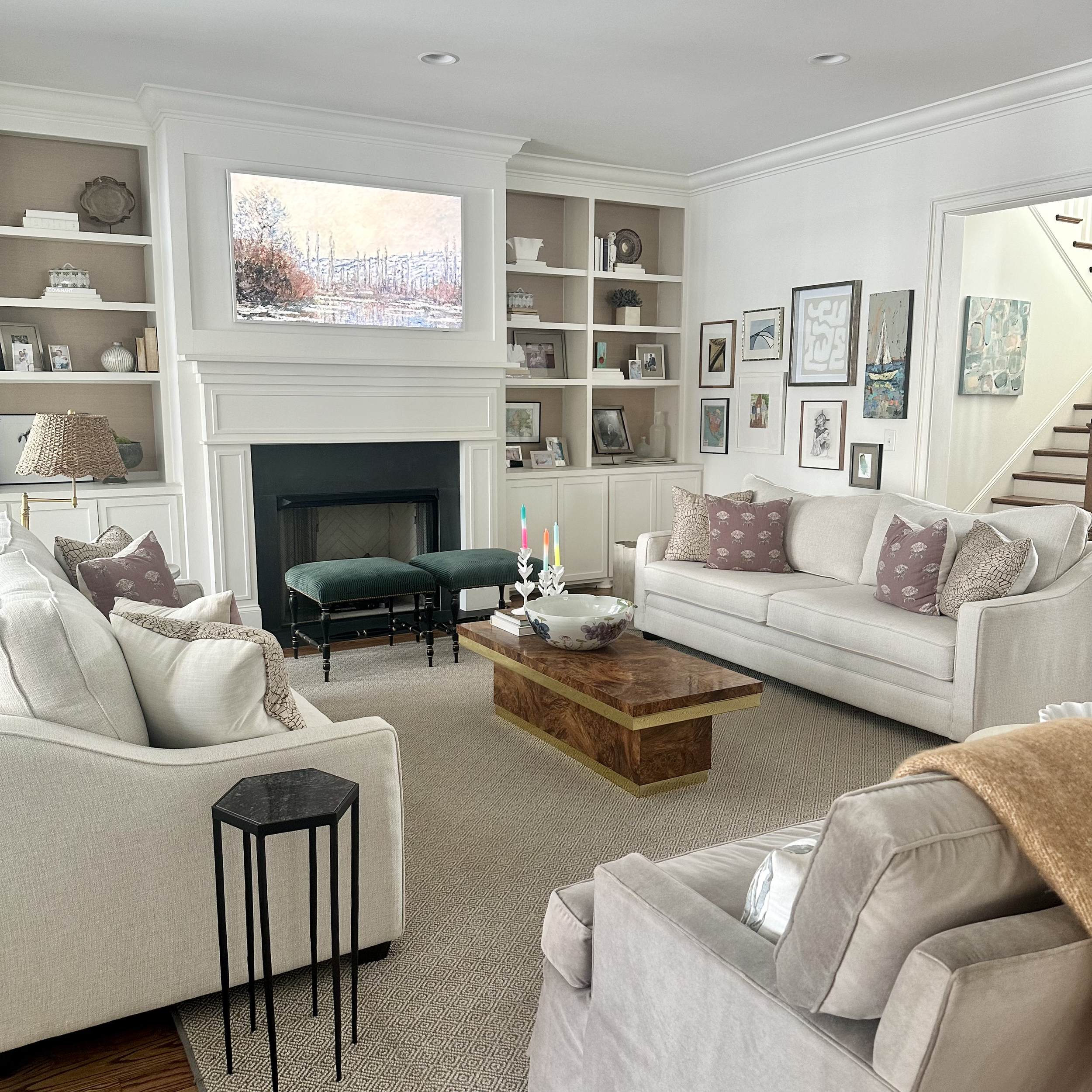 Cozy living room with a modern design, featuring light-colored sofas, patterned throw pillows, a wooden coffee table, and a fireplace with a mantle. The room is decorated with framed artwork on the walls and built-in shelves displaying decor items. T