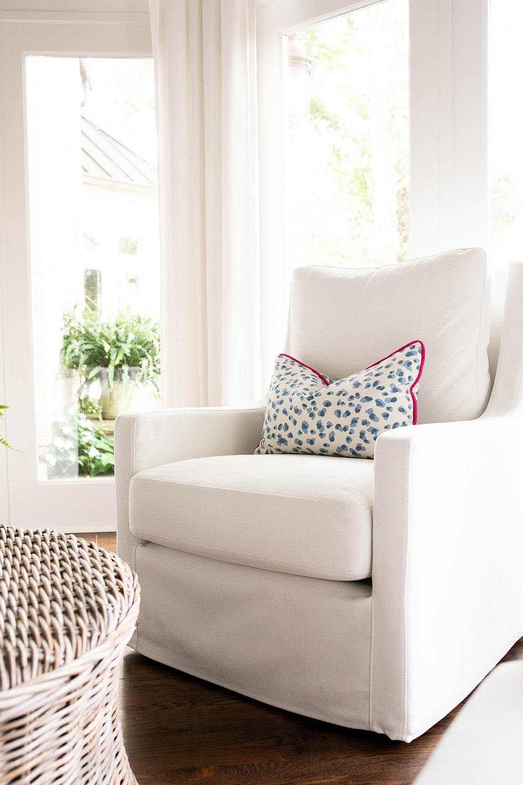 White armchair with decorative pillow, wicker table, large windows with sheer curtains, and houseplants in the background.