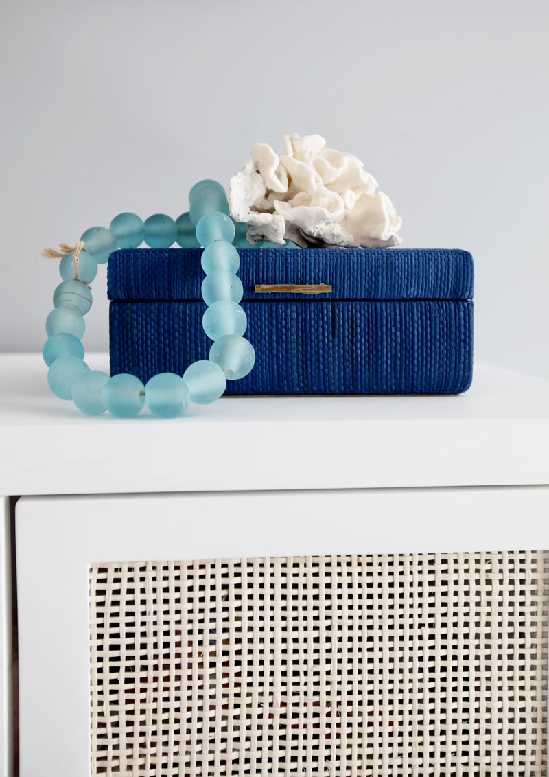 Blue jewelry box with white coral and blue beads on top, placed on a white shelf.