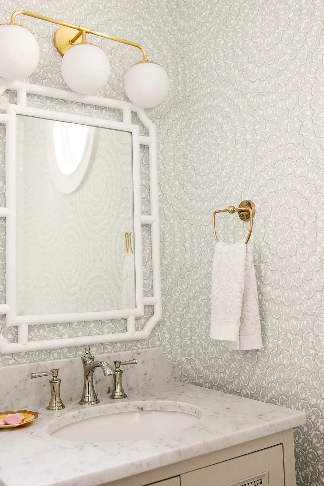 Elegant bathroom with patterned wallpaper, white-framed mirror, brass light fixture, marble countertop, silver faucet, and white towel on a gold holder.