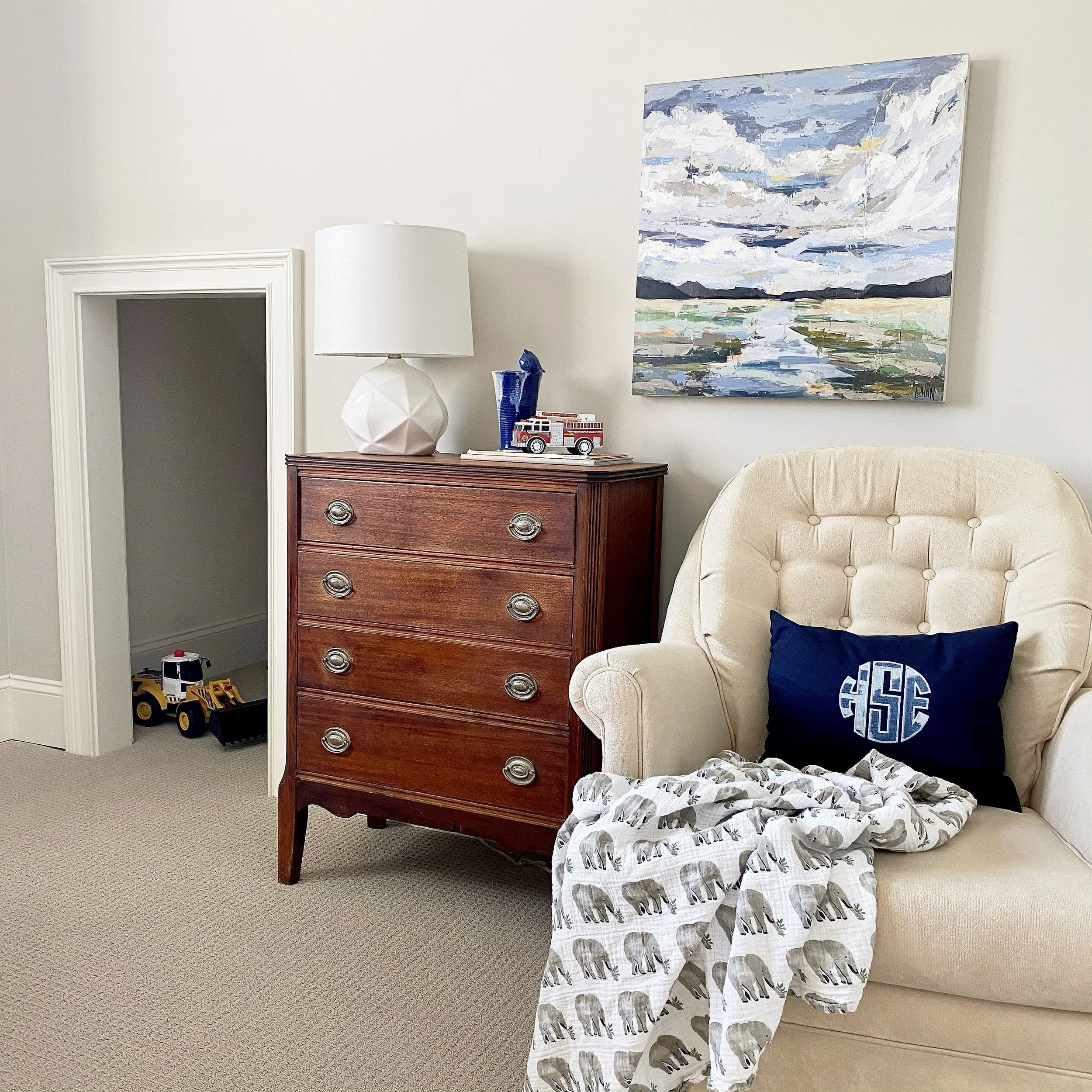 Cozy room with a beige armchair, a wooden dresser with a lamp and toy fire truck, abstract landscape painting, and toy bulldozer.
