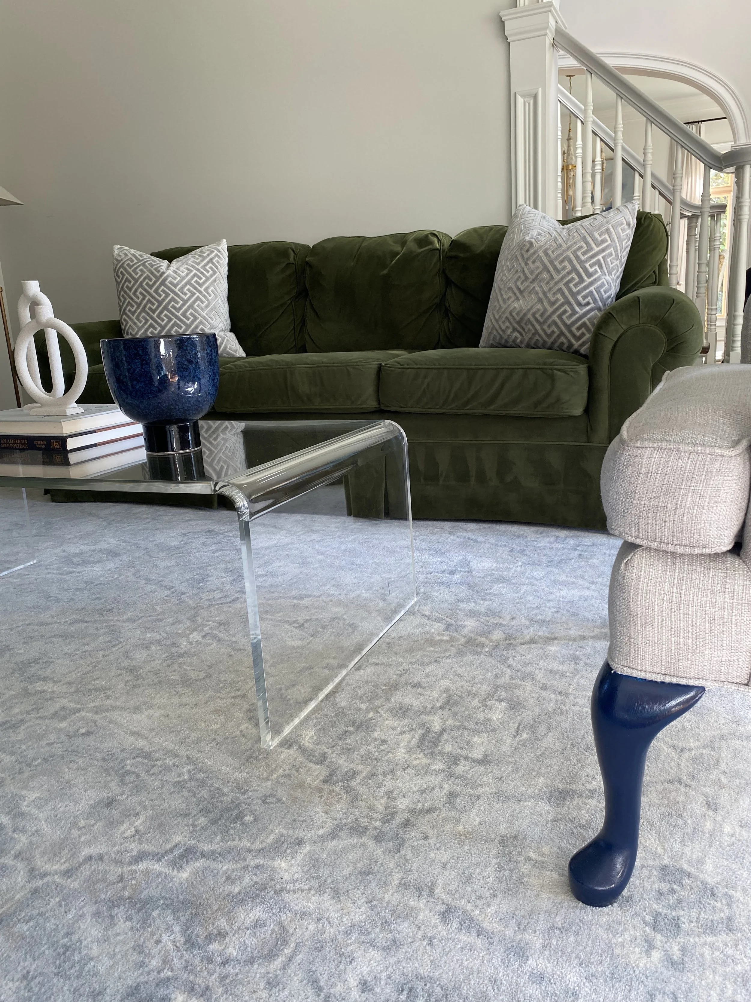 Living room with green velvet sofa, geometric pillows, clear glass coffee table, gray rug, and blue legged armchair.