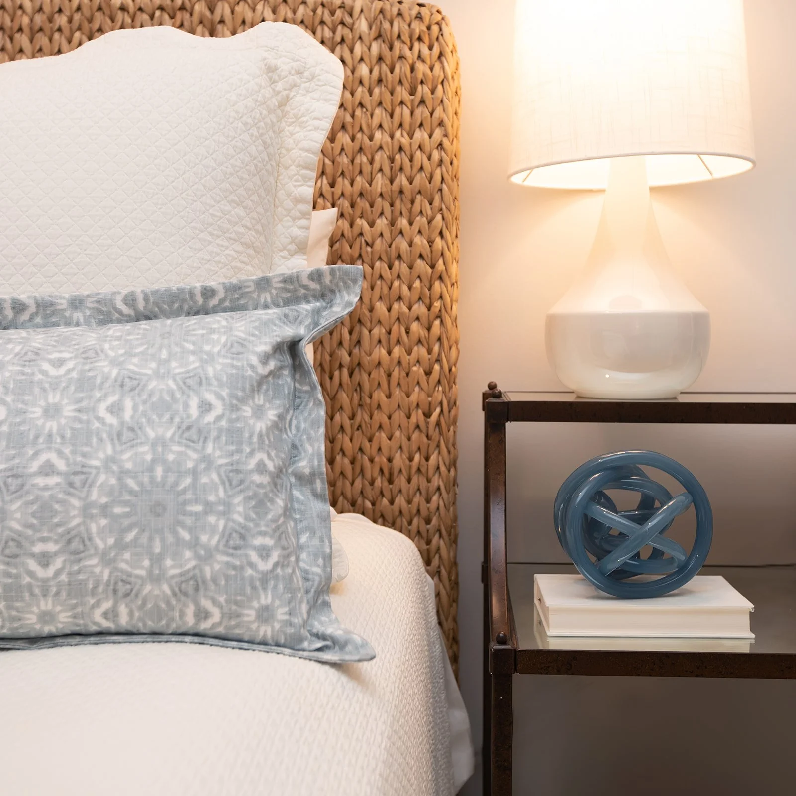 Bedroom scene with woven headboard, patterned blue pillow, white quilt, bedside table, white lamp, and abstract blue sculpture.