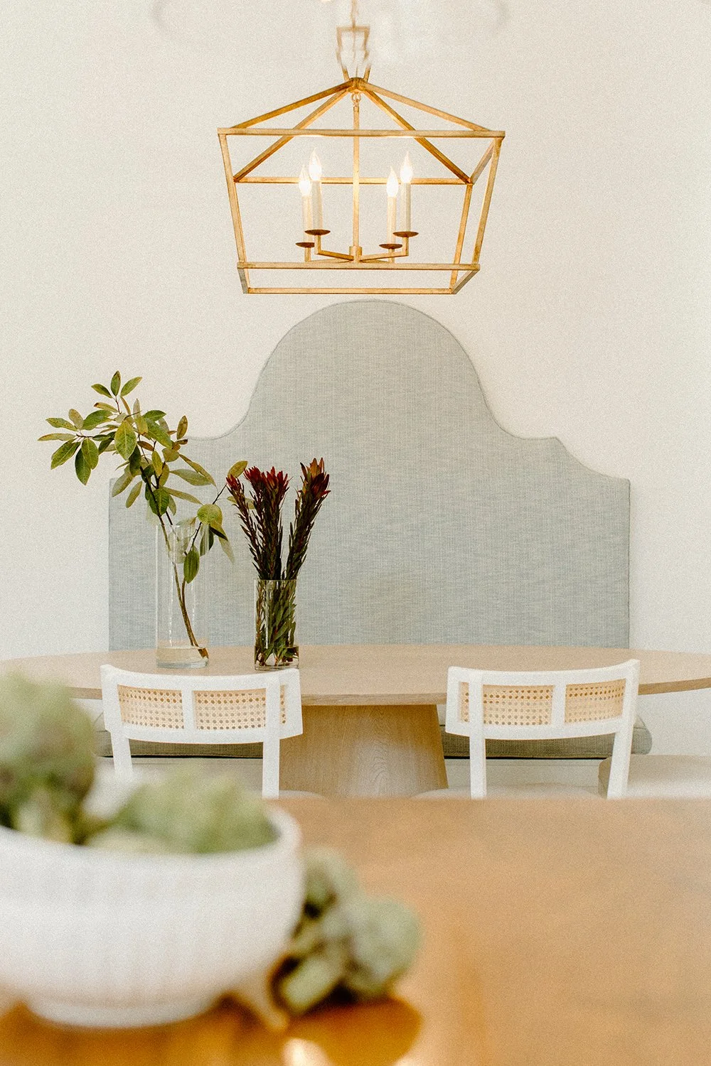 A dining area with a light blue upholstered bench, a round wooden table, white chairs with cane backs, a gold geometric chandelier, and vases with green and red foliage.