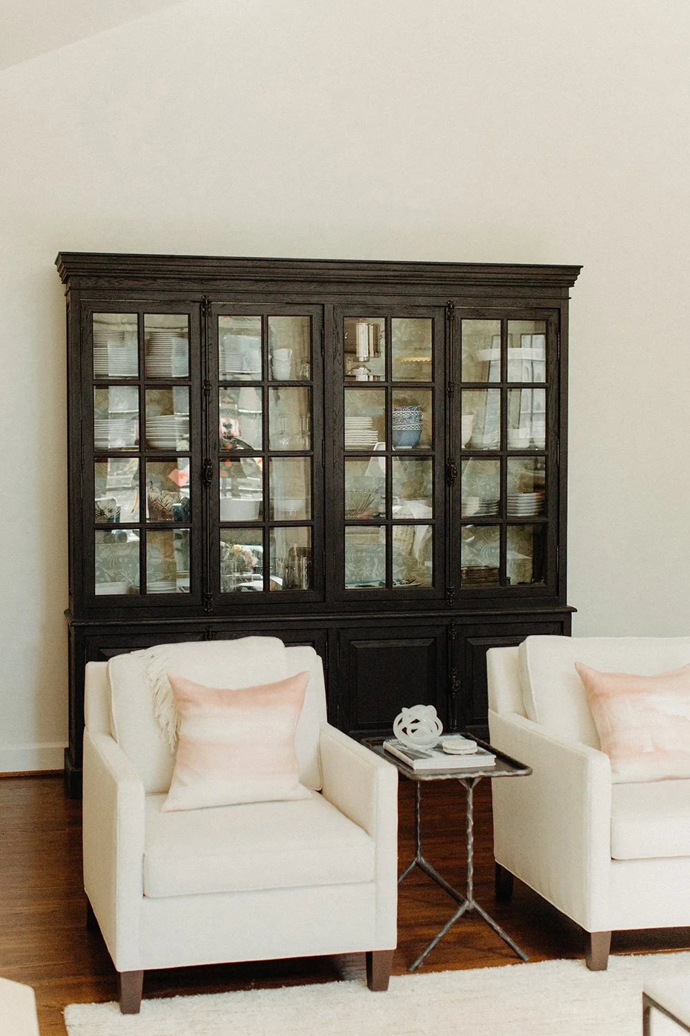 Living room with black china cabinet, two white armchairs with pink pillows, and a small side table on a hardwood floor.