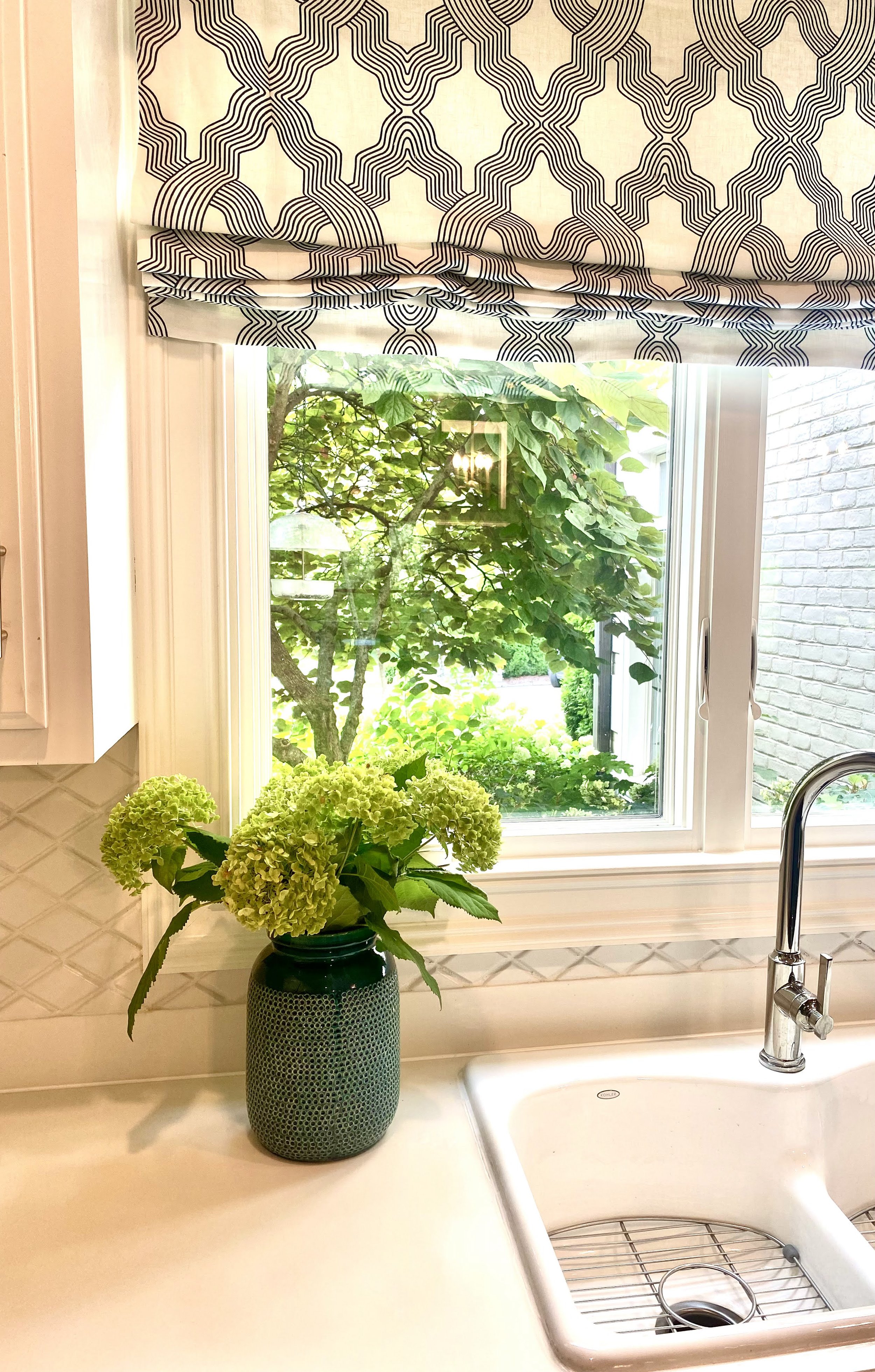 A kitchen window with a patterned Roman shade, a green vase with hydrangeas on the counter, and a white sink with a silver faucet.