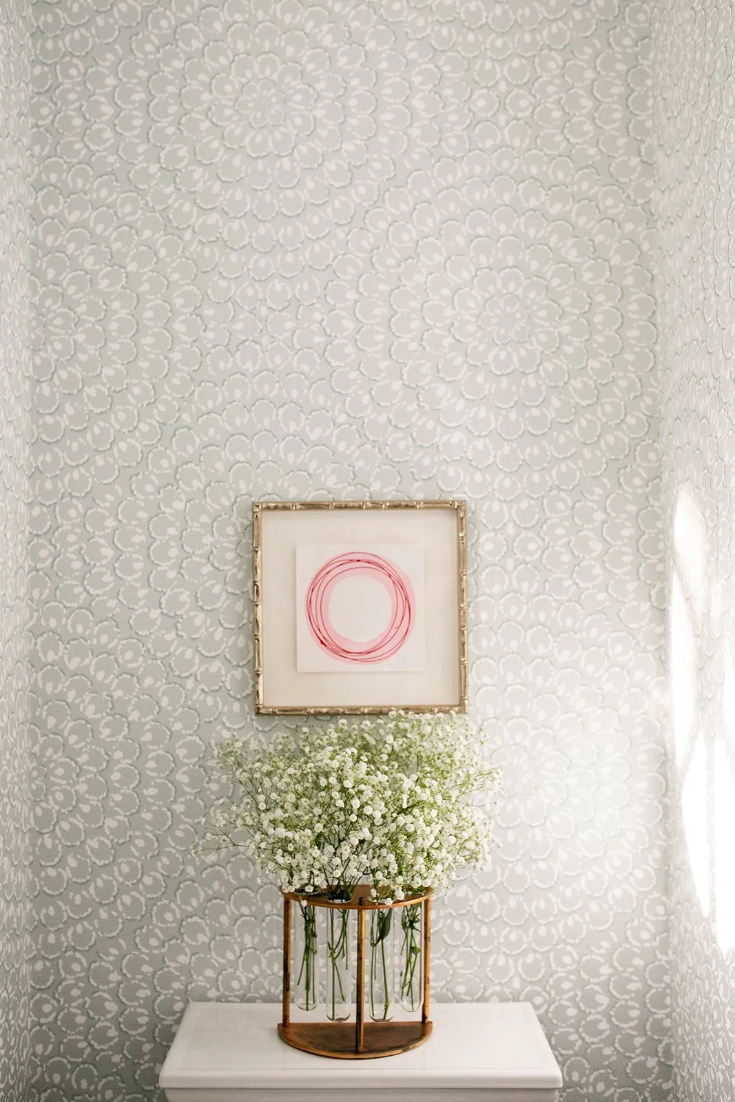 Decorative room corner with floral patterned wallpaper, a framed red circular art piece above, and a vase with baby's breath flowers on a white surface.